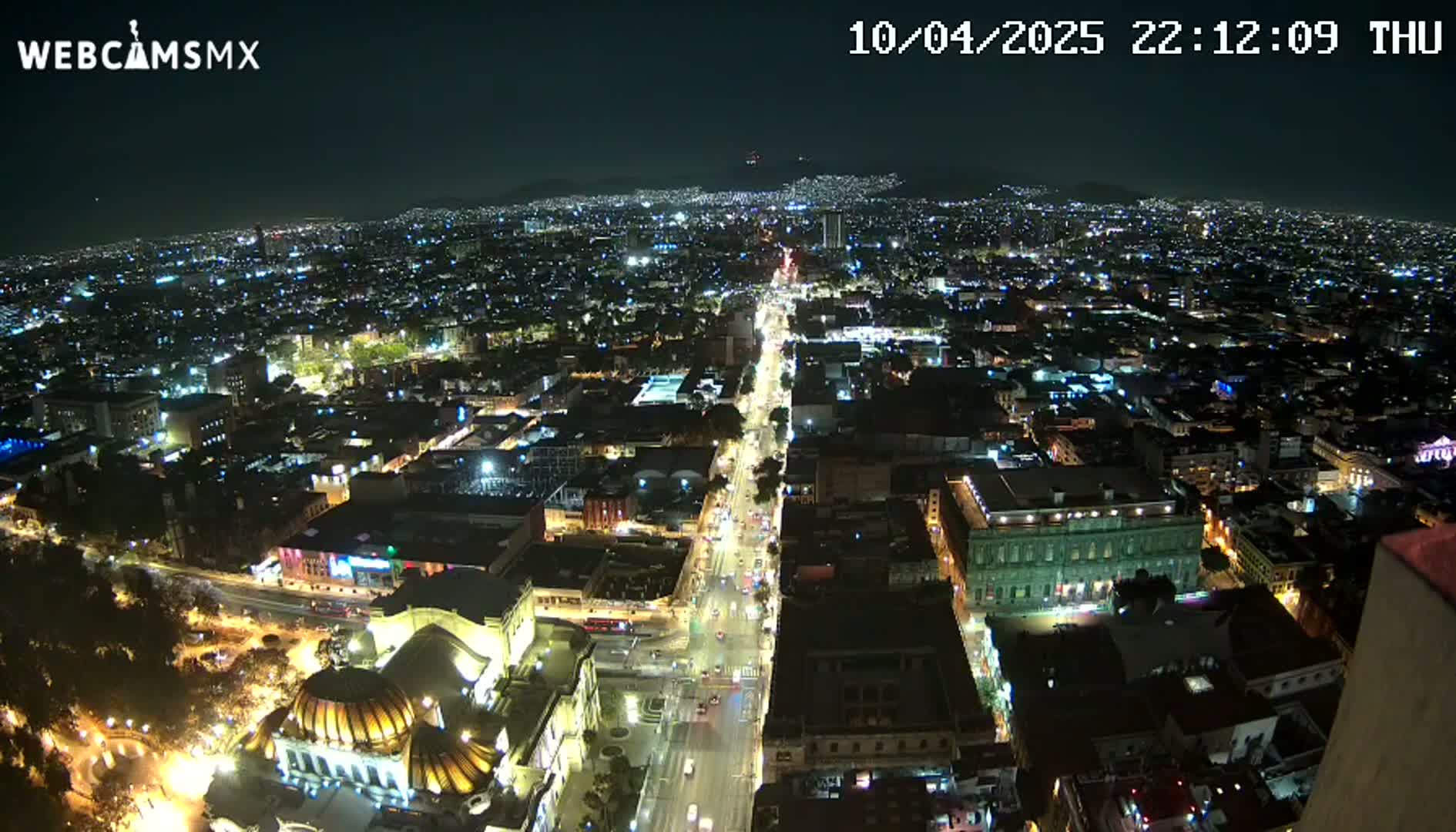 A nighttime aerial view of a brightly lit city, showing a long, illuminated street stretching towards distant mountains under a clear sky.