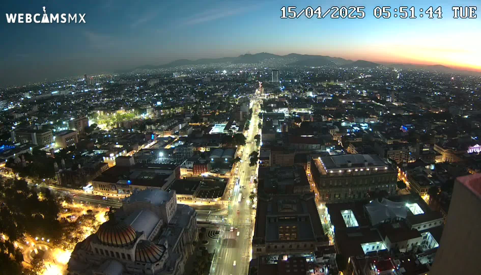 An aerial nighttime view of a city with illuminated streets and buildings, under a twilight sky with distant mountains visible on the horizon.