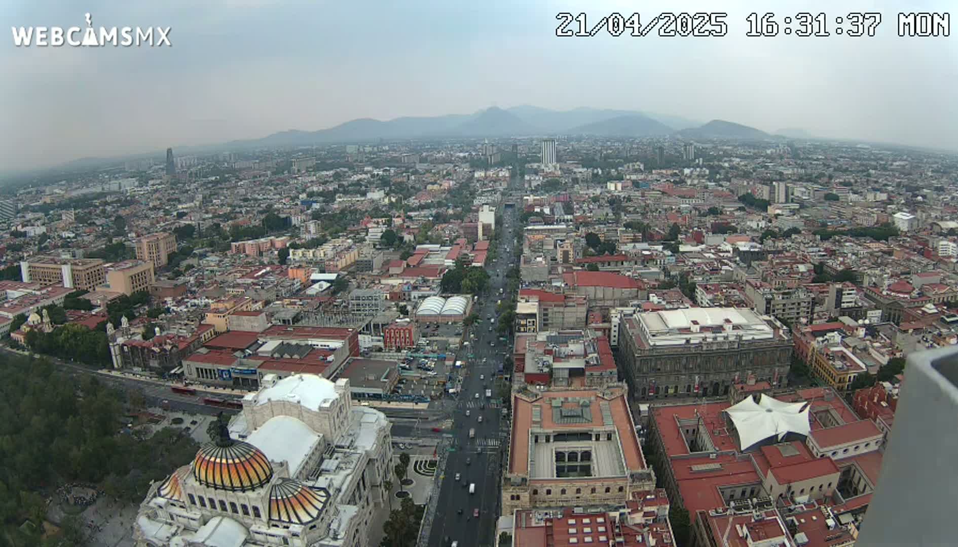 A hazy, high-angle view shows a vast cityscape with a long, wide avenue cutting through it, culminating in a large domed building at the bottom; mountains are visible in the distance.