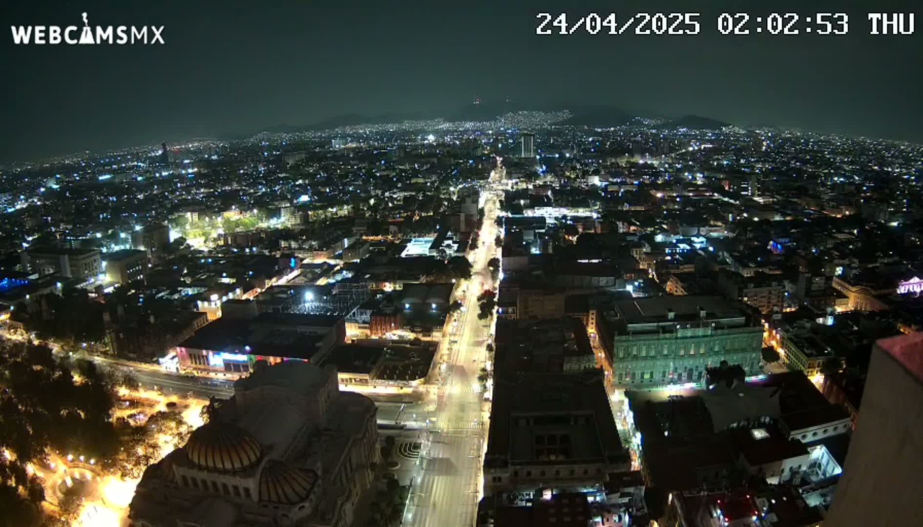 A nighttime, high-angle view of a sprawling city illuminated by streetlights and building lights, with dark mountains in the background under a clear sky.