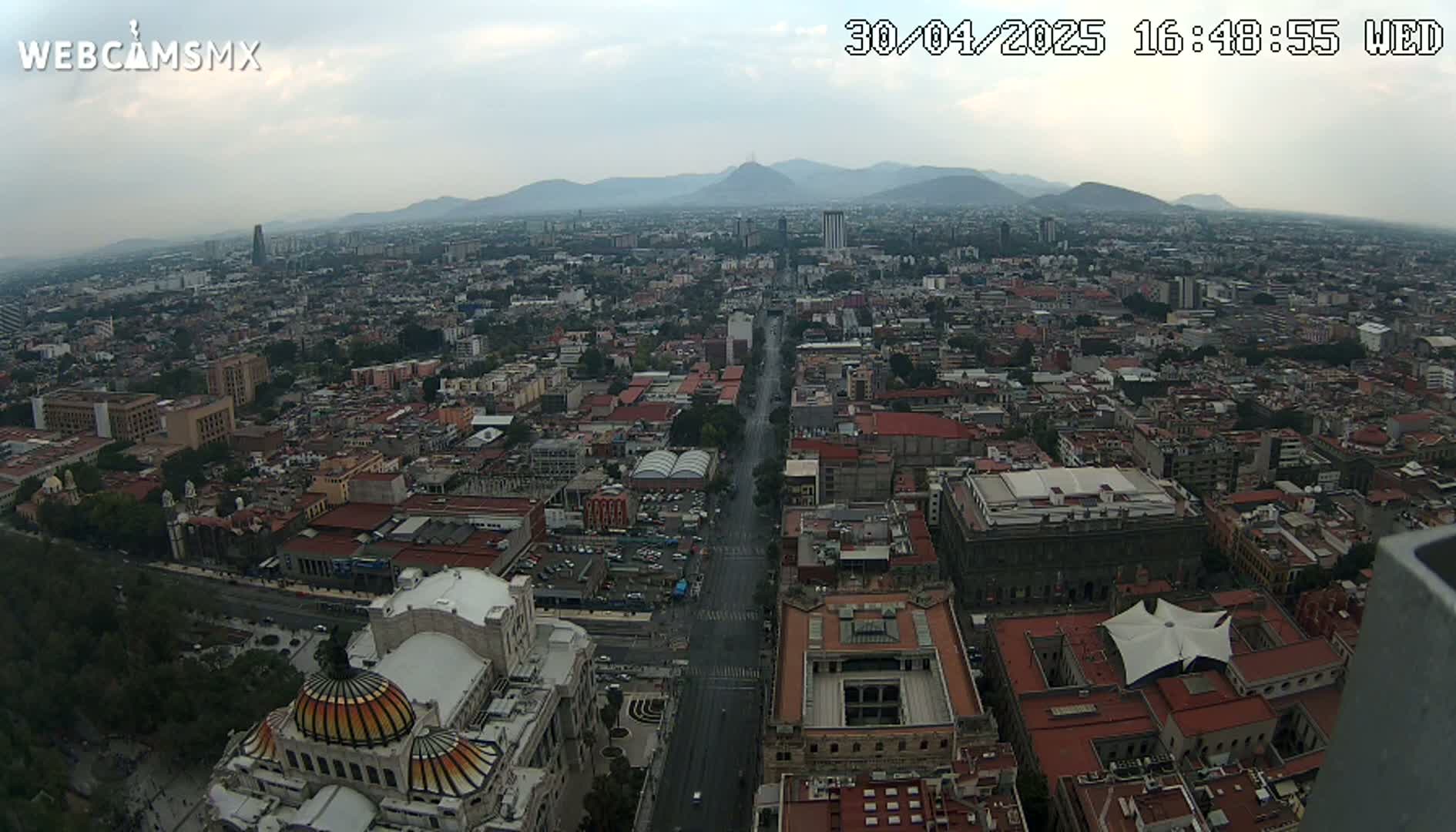 A high-angle, long shot of a city on a somewhat overcast day, showing a wide, long avenue cutting through a dense urban landscape with mountains visible in the distance.