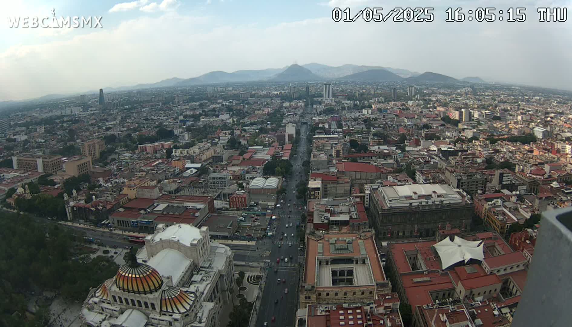 A hazy, daytime, high-angle view of a sprawling city with a long, straight road running through its center, mountains visible in the background.