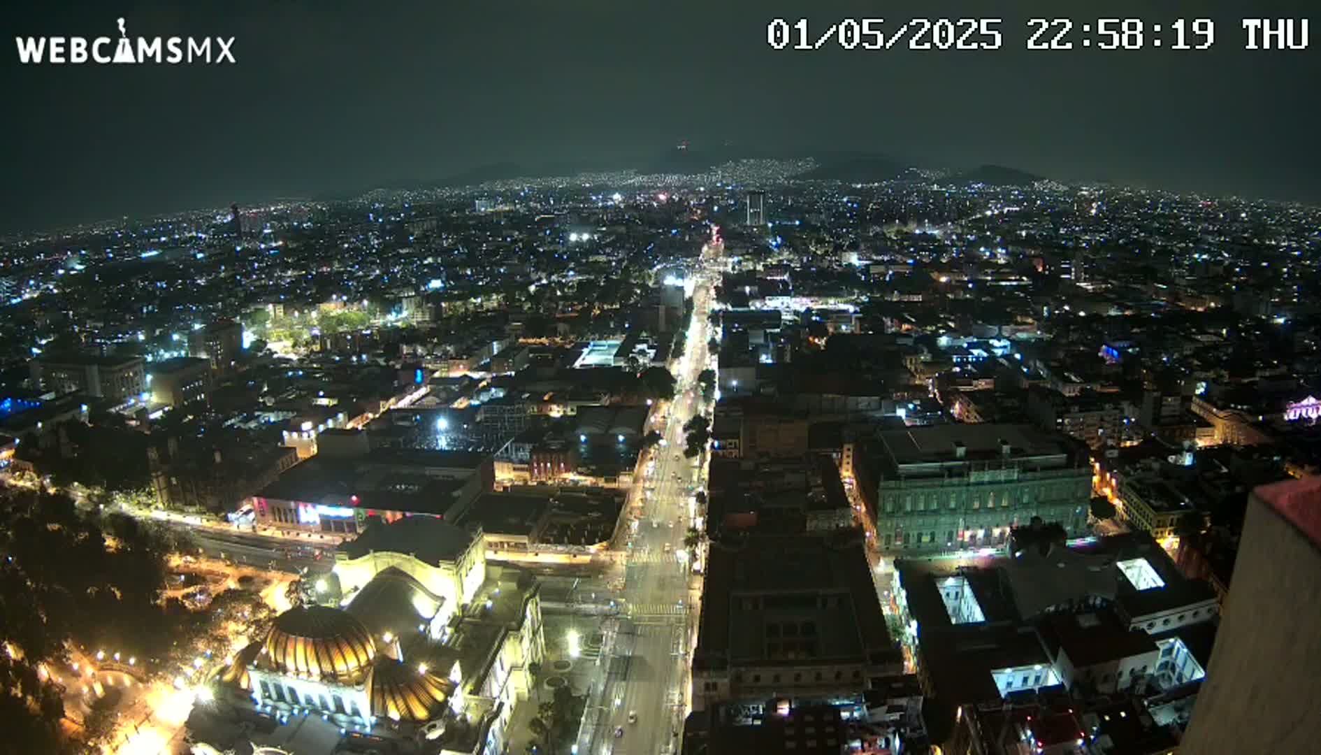 A nighttime aerial view shows a sprawling city illuminated by streetlights and building lights, with a long, brightly lit avenue cutting through the urban landscape.