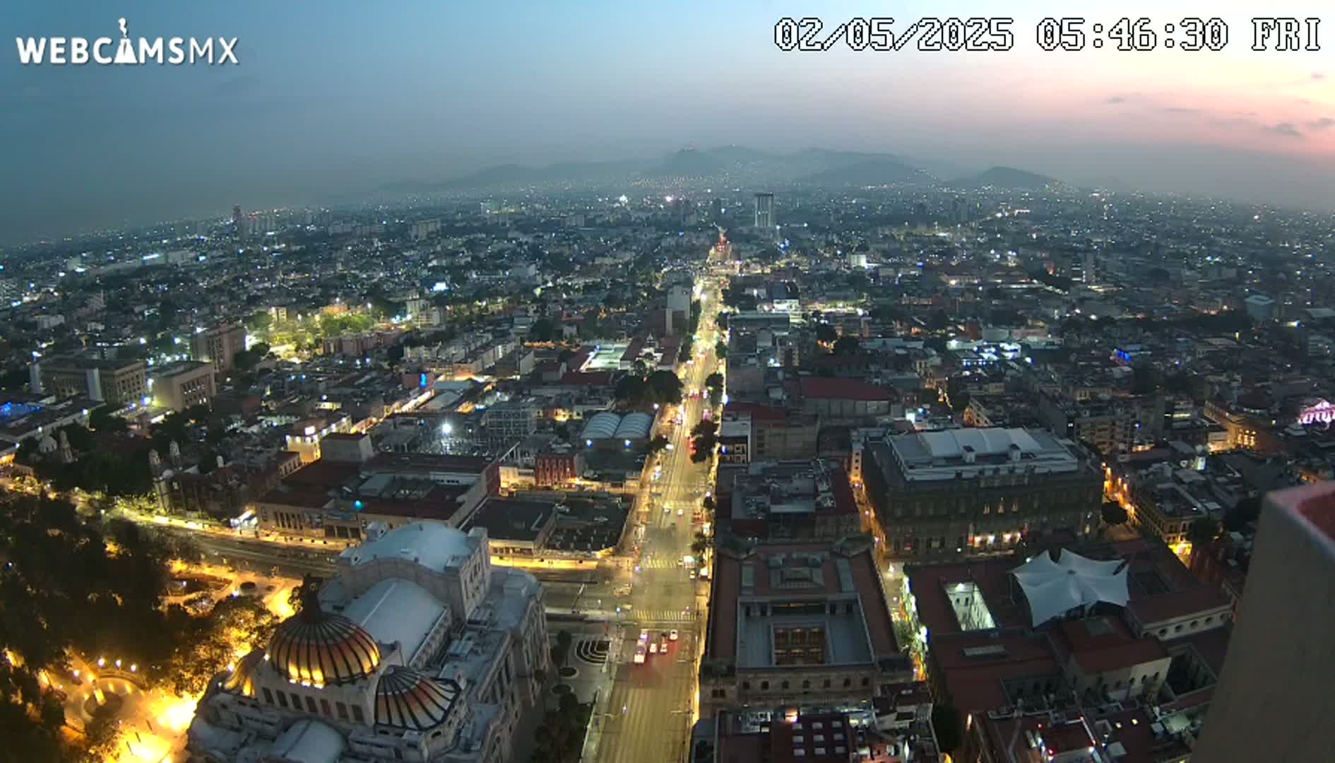 A nighttime, high-angle view of a sprawling city illuminated by streetlights and building lights, with mountains visible in the distance under a twilight sky.
