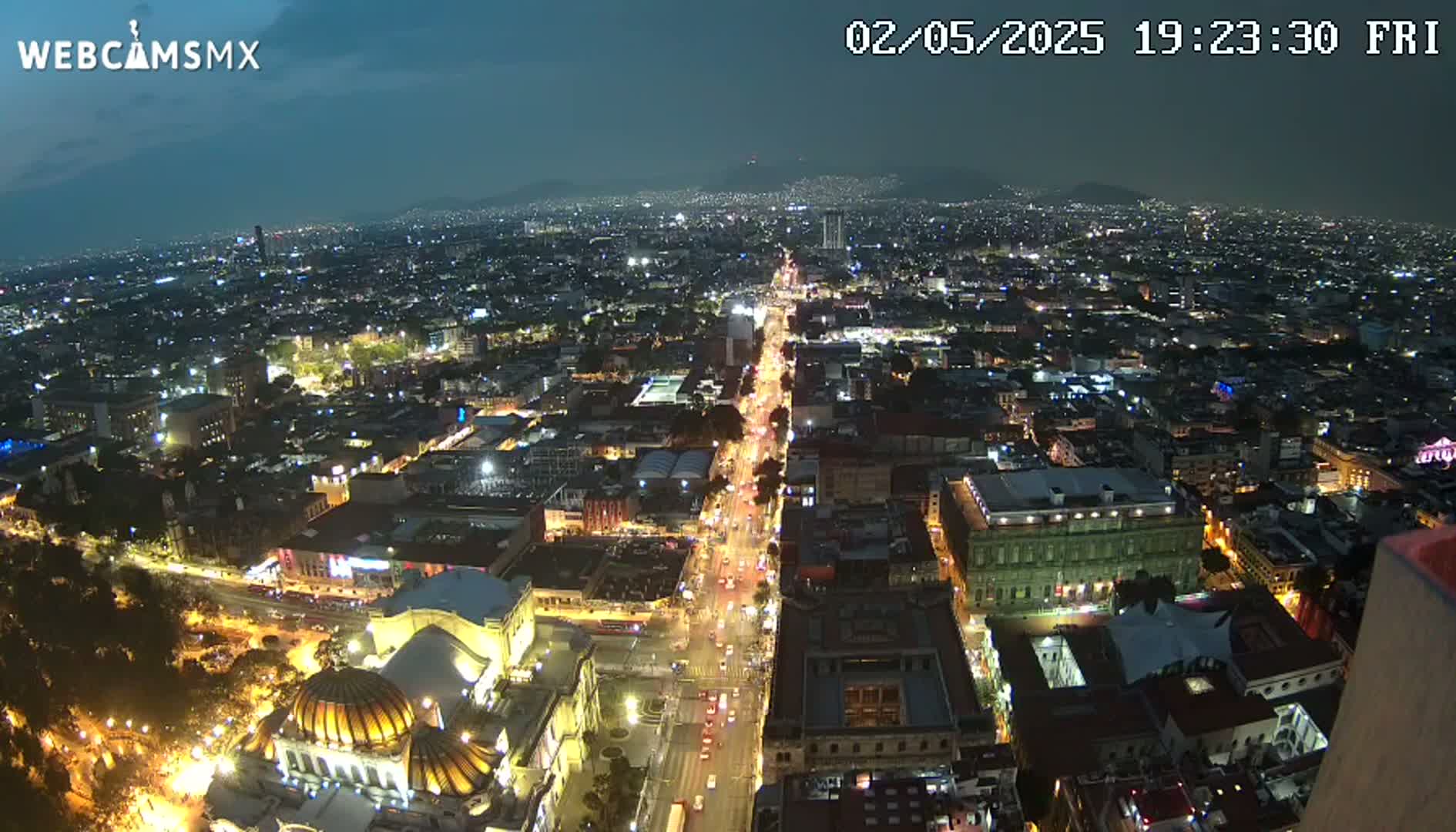 A nighttime, high-angle view of a brightly lit city, with a long, illuminated street stretching toward distant mountains under a dark sky.