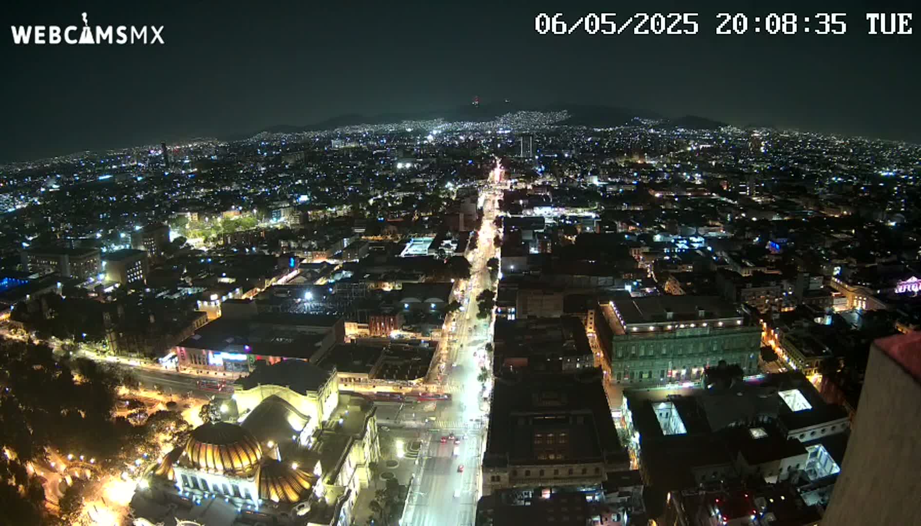 A nighttime, high-angle view shows a sprawling city illuminated by streetlights and building lights, with mountains visible in the distance under a clear sky.