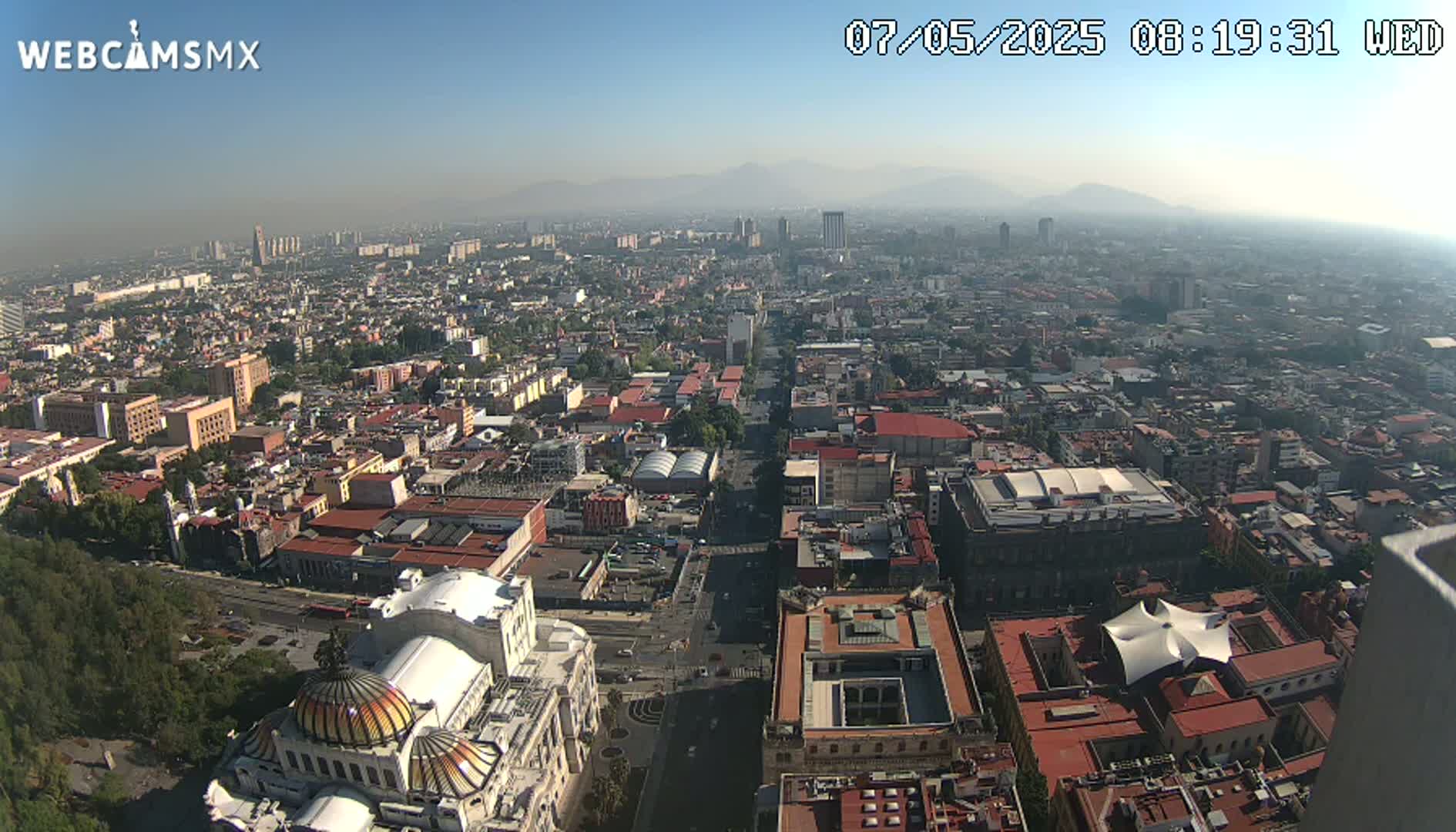 A hazy, sunny day reveals a sprawling cityscape with a large, ornate building in the foreground, mountains visible in the distance.