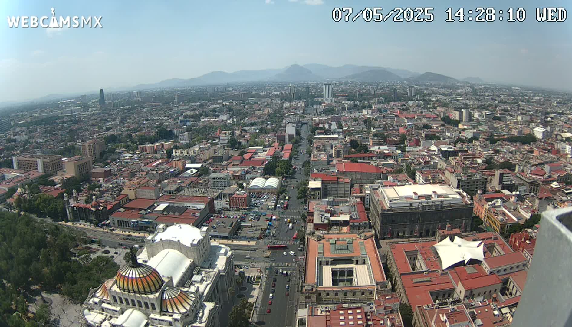 A high-angle, long shot on a hazy day shows a sprawling city with red-tiled roofs, a wide avenue, and distant mountains under a light-blue sky.