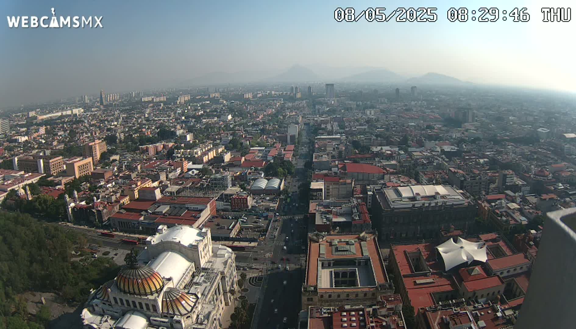 A hazy, high-angle view of a sprawling city, showing a large building with a distinctive dome in the foreground, mountains in the distance.