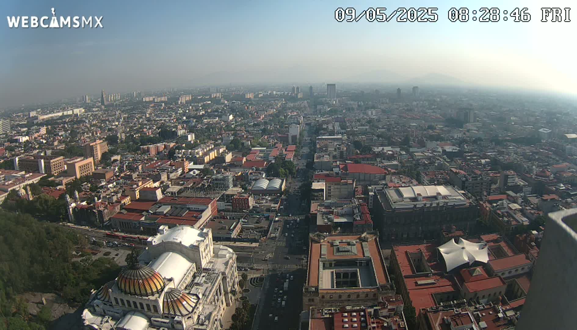 A hazy, high-angle view shows a sprawling city with red-tiled roofs, a large building with a dome, and a long, straight road cutting through the urban landscape.