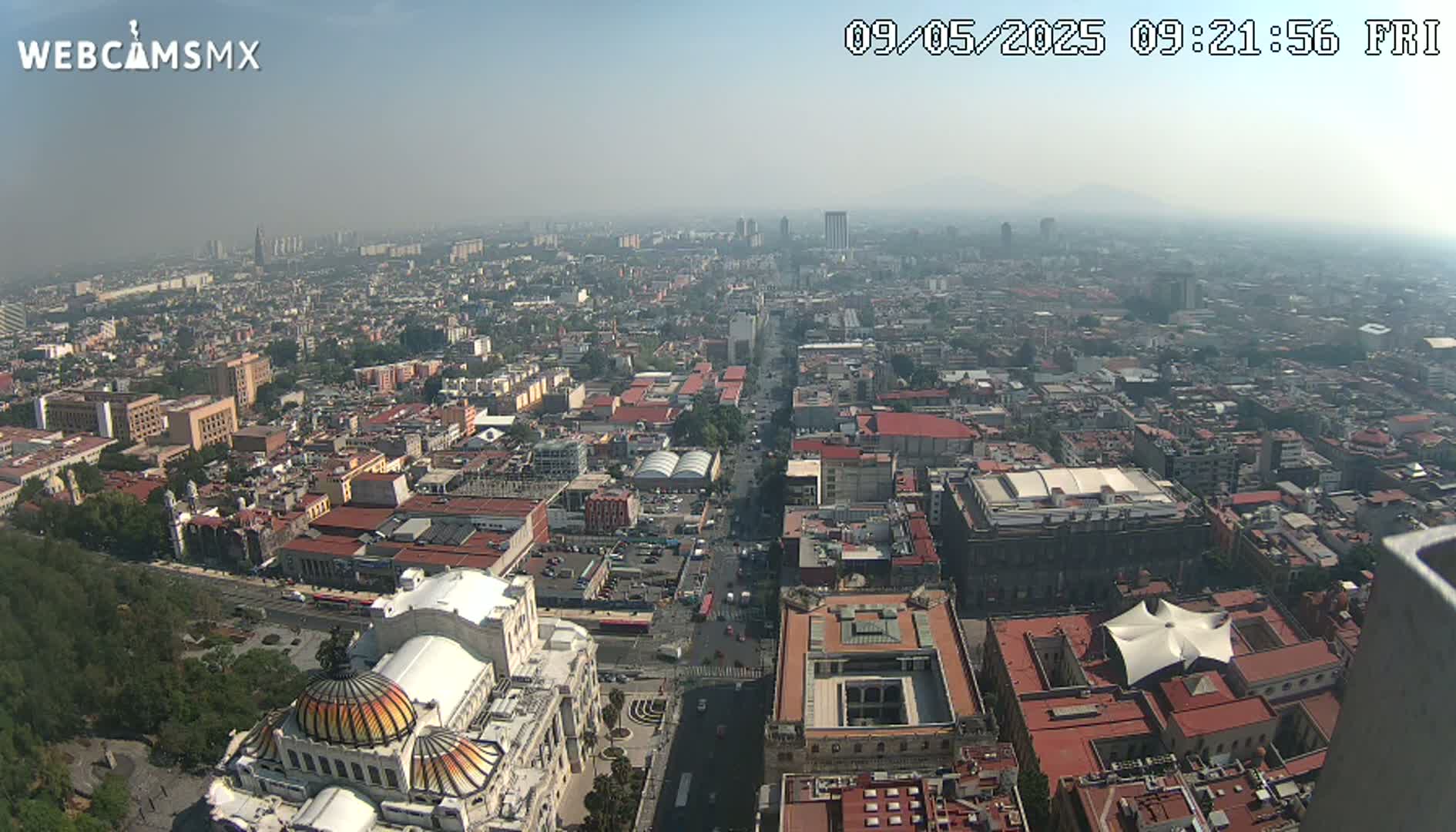 A hazy, high-angle view shows a sprawling city with red-tiled roofs, a large building with a dome, and a wide street stretching into the distance.