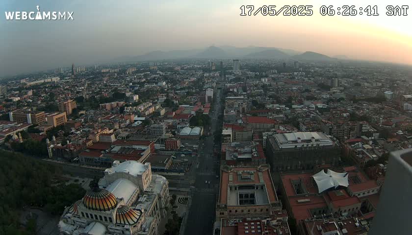 A hazy sunset illuminates a sprawling cityscape, viewed from a high vantage point, showing a long avenue cutting through the city toward distant mountains.