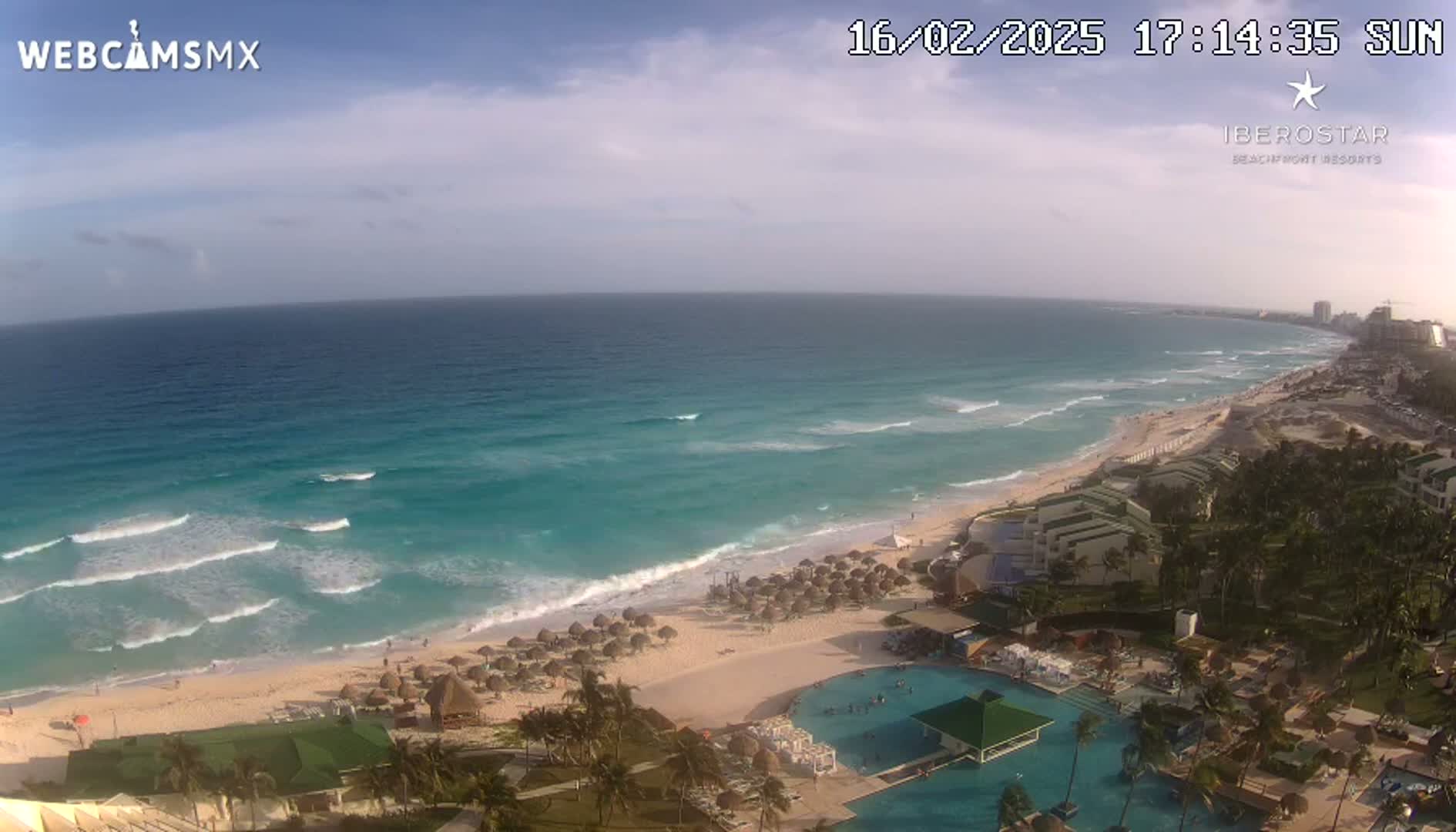 An aerial view of a beach resort on a sunny day shows turquoise water with waves lapping a sandy shore lined with beach umbrellas, palm trees, and resort buildings with a large pool.