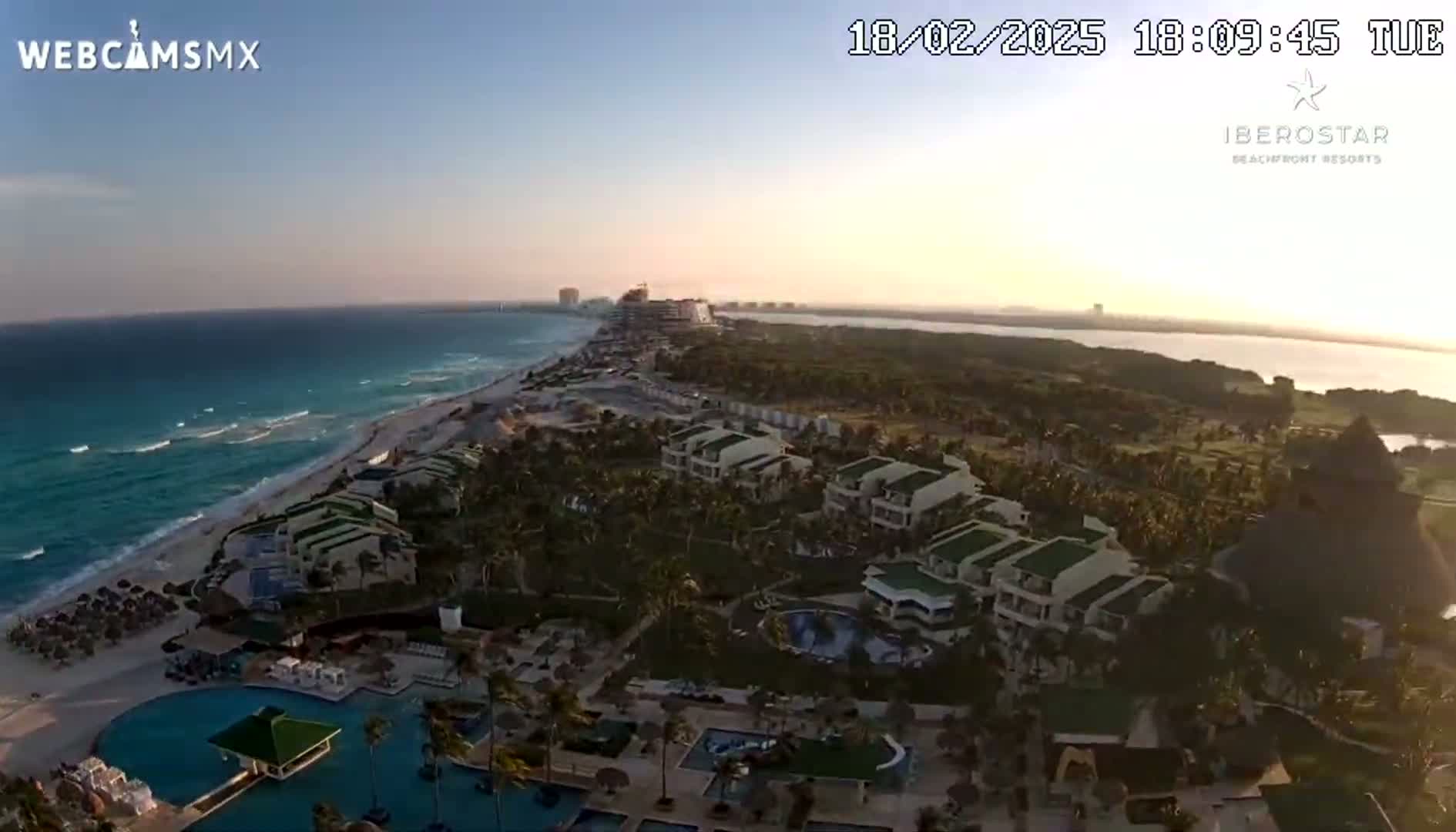 An aerial view shows a beach resort with pools and buildings situated along a sandy beach next to a calm ocean at sunset.