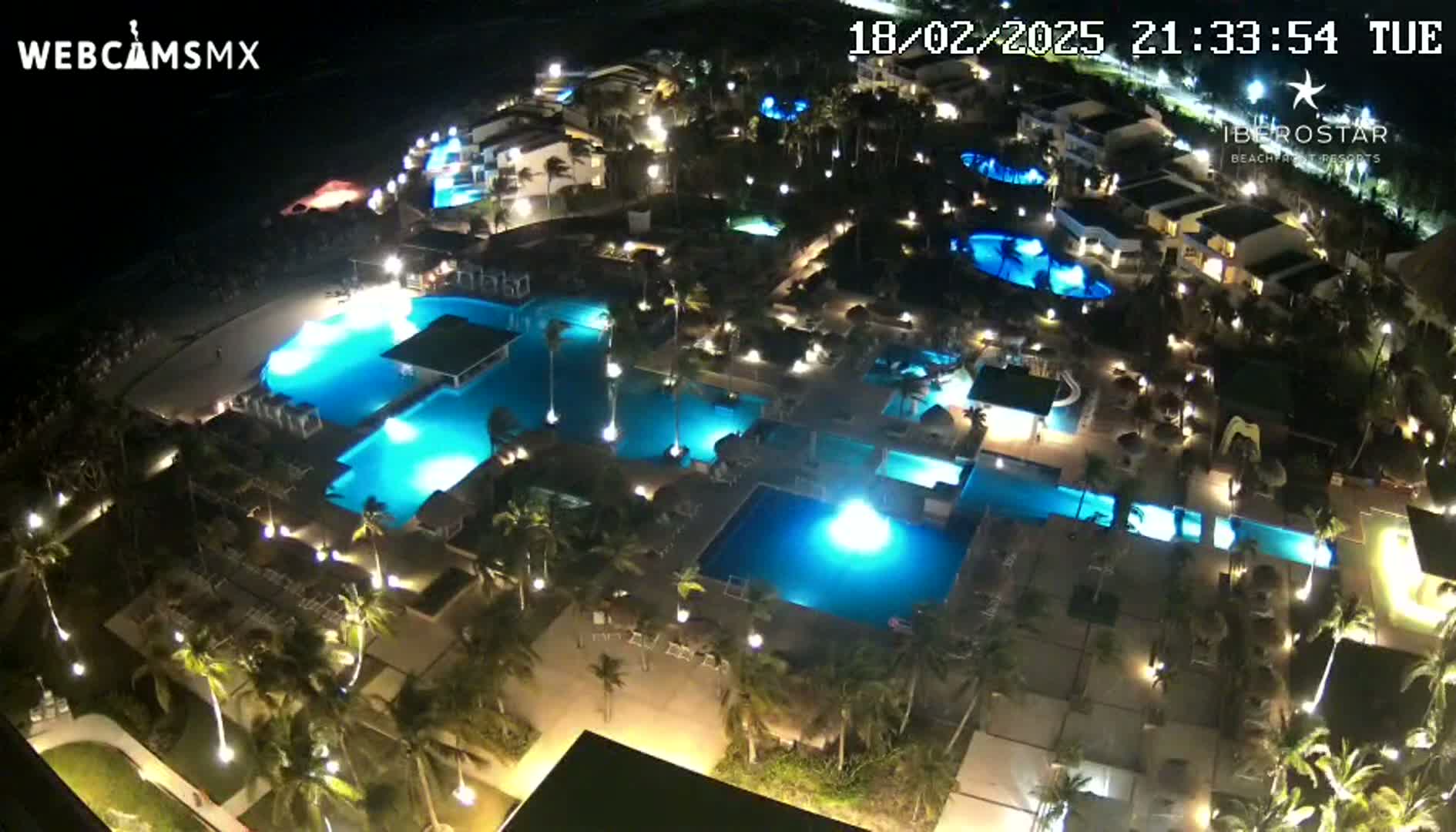 An aerial nighttime view shows a resort complex with numerous illuminated swimming pools surrounded by palm trees and buildings.