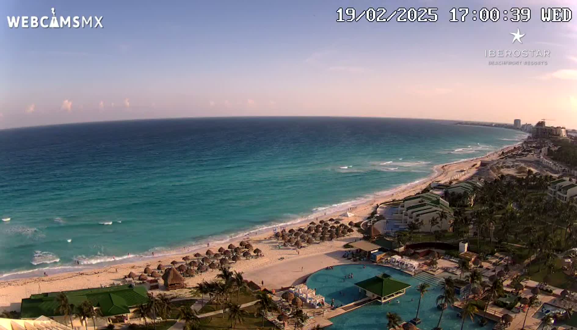 A beach resort with turquoise water and a sandy beach is seen under a clear, sunset sky.