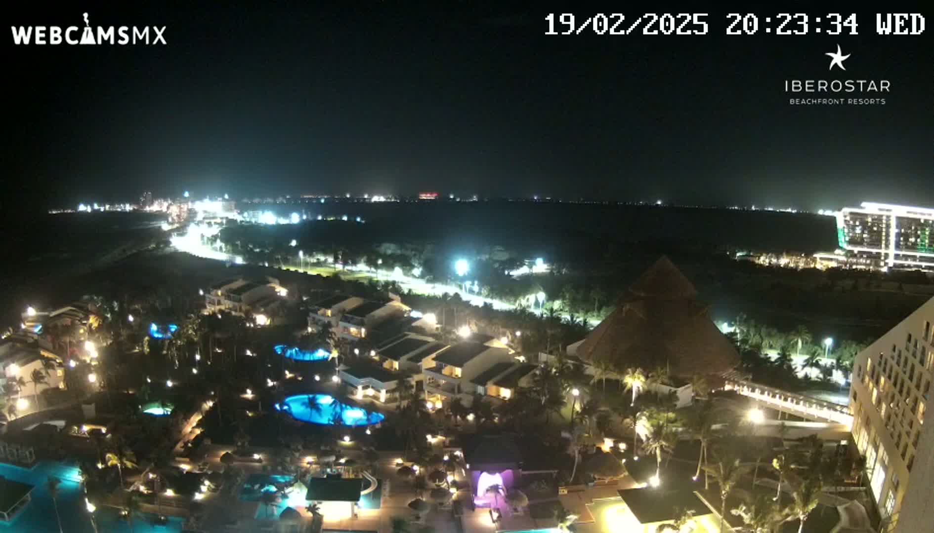 A nighttime, aerial view of a resort complex with illuminated buildings, pools, and landscaping, extending to a coastline with city lights in the distance under a clear sky.