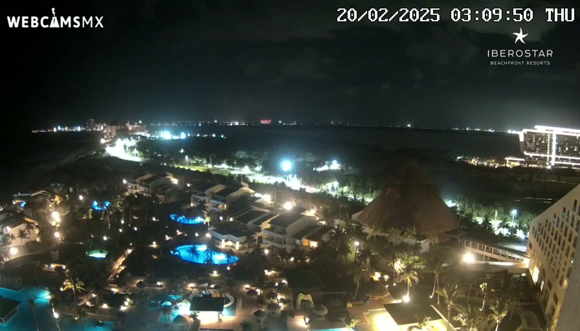A nighttime aerial view of a resort complex with illuminated buildings, pools, and landscaping, overlooking a calm body of water and a distant city skyline under a clear sky.