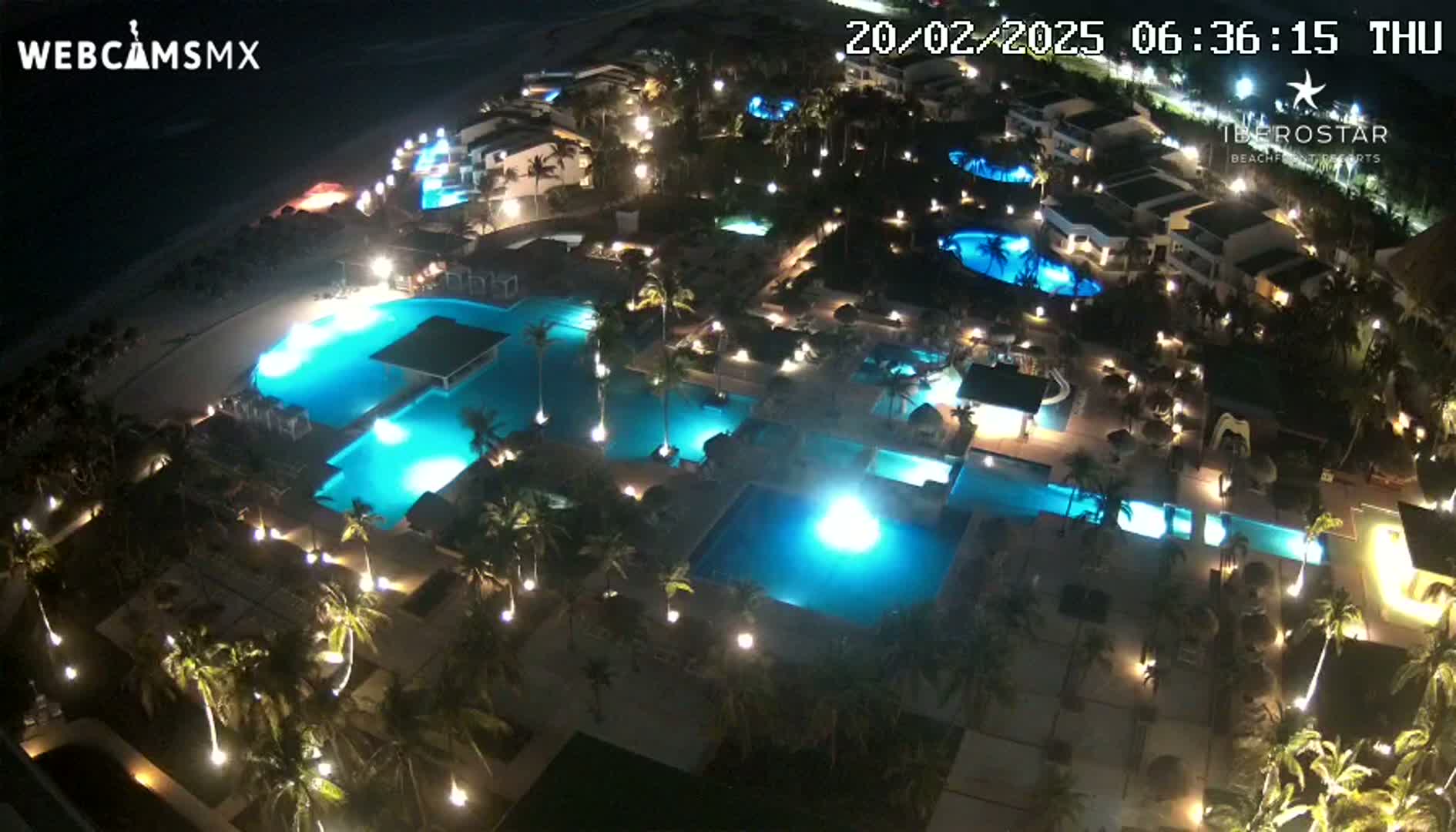 An illuminated resort at night, featuring numerous pools and palm trees, sits on a beach under a dark sky.
