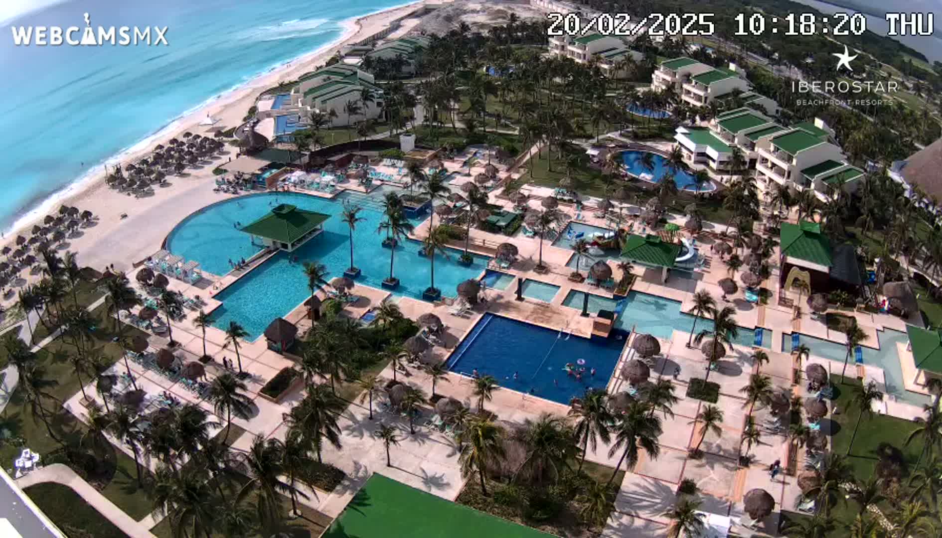 An aerial view shows a resort with multiple pools, surrounding buildings, palm trees, and a beach with turquoise water under sunny skies.