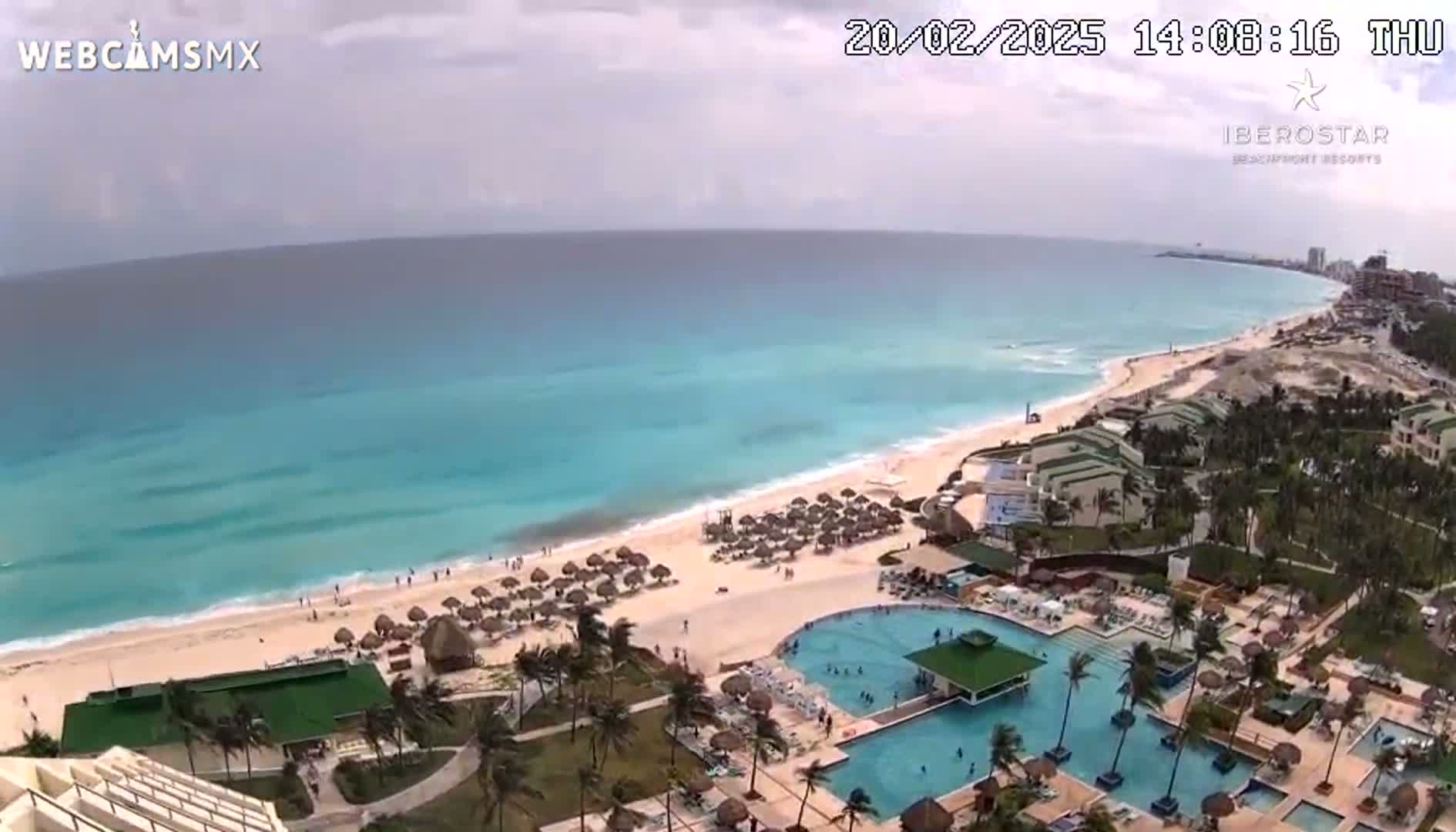 An aerial view shows a beach resort with multiple pools and palm trees under partly cloudy skies, next to a long, light-blue ocean with small waves.
