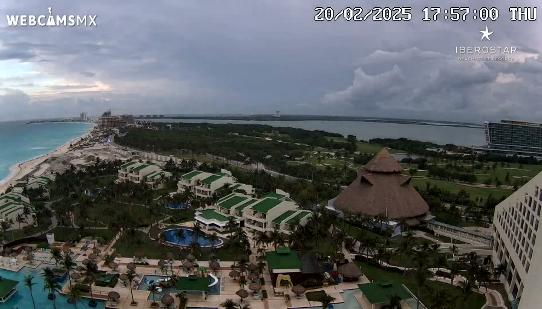An aerial view shows a resort complex with pools and buildings under a cloudy sky, overlooking a beach and ocean.