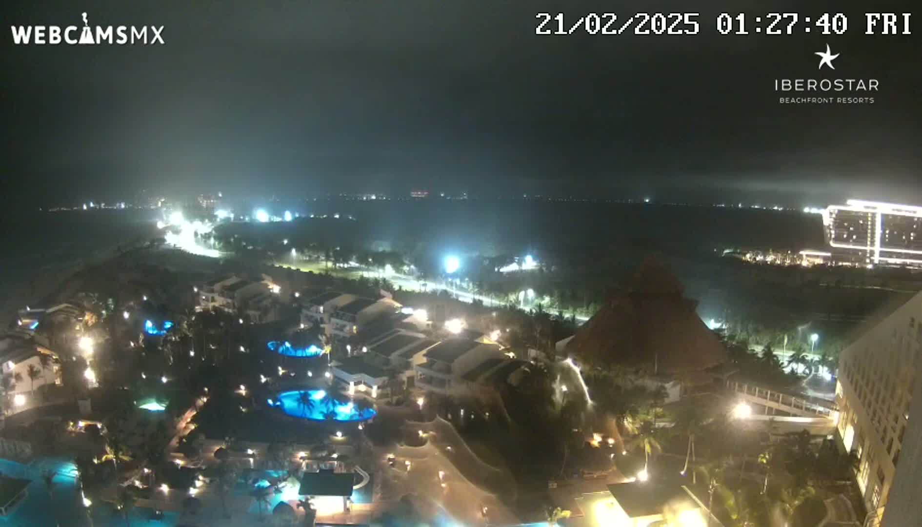 A nighttime aerial view of a resort complex with pools and buildings illuminated, overlooking a dark ocean under hazy conditions.