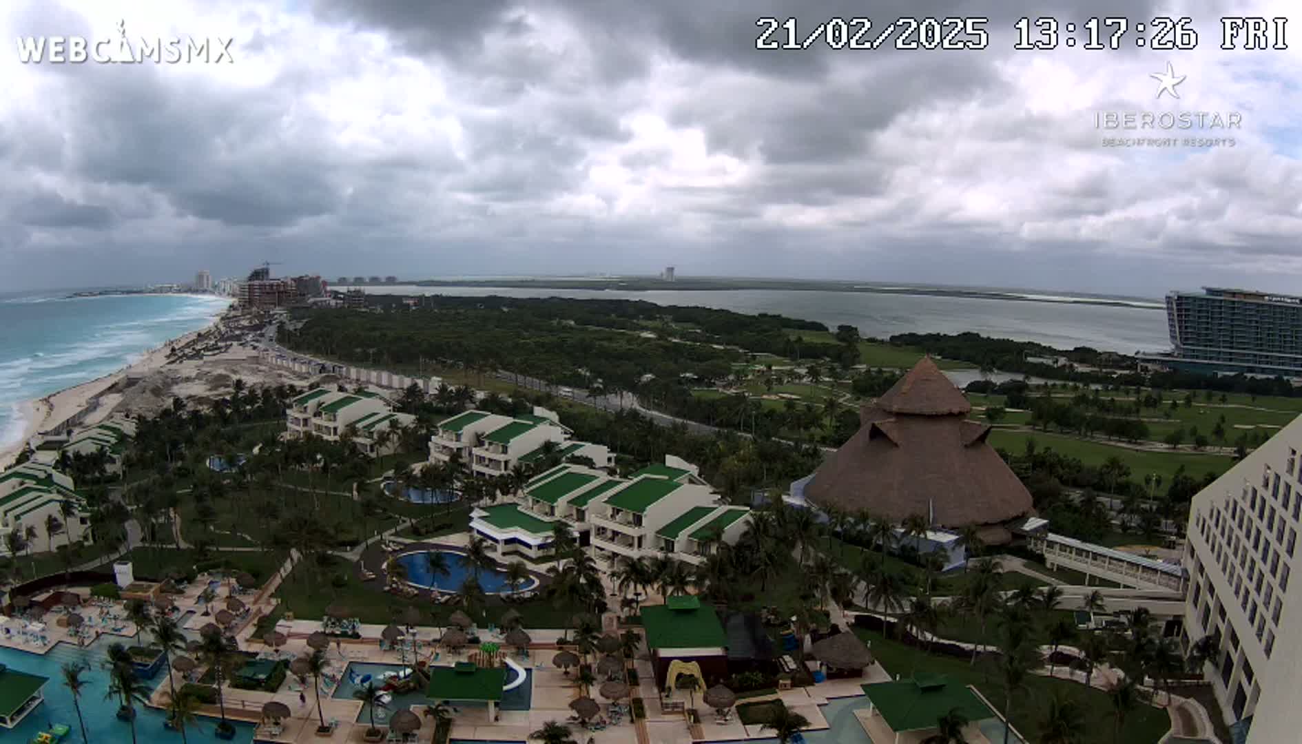 An aerial view shows a resort area with pools, buildings, and lush landscaping, overlooking a beach and ocean under a cloudy sky.