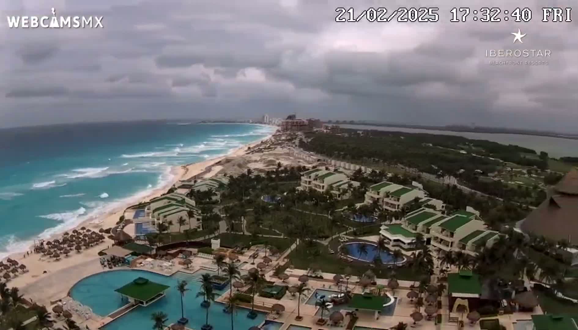 An overcast sky hangs over a resort town on a beach with turquoise water and white waves, featuring numerous buildings with pools and lush landscaping.