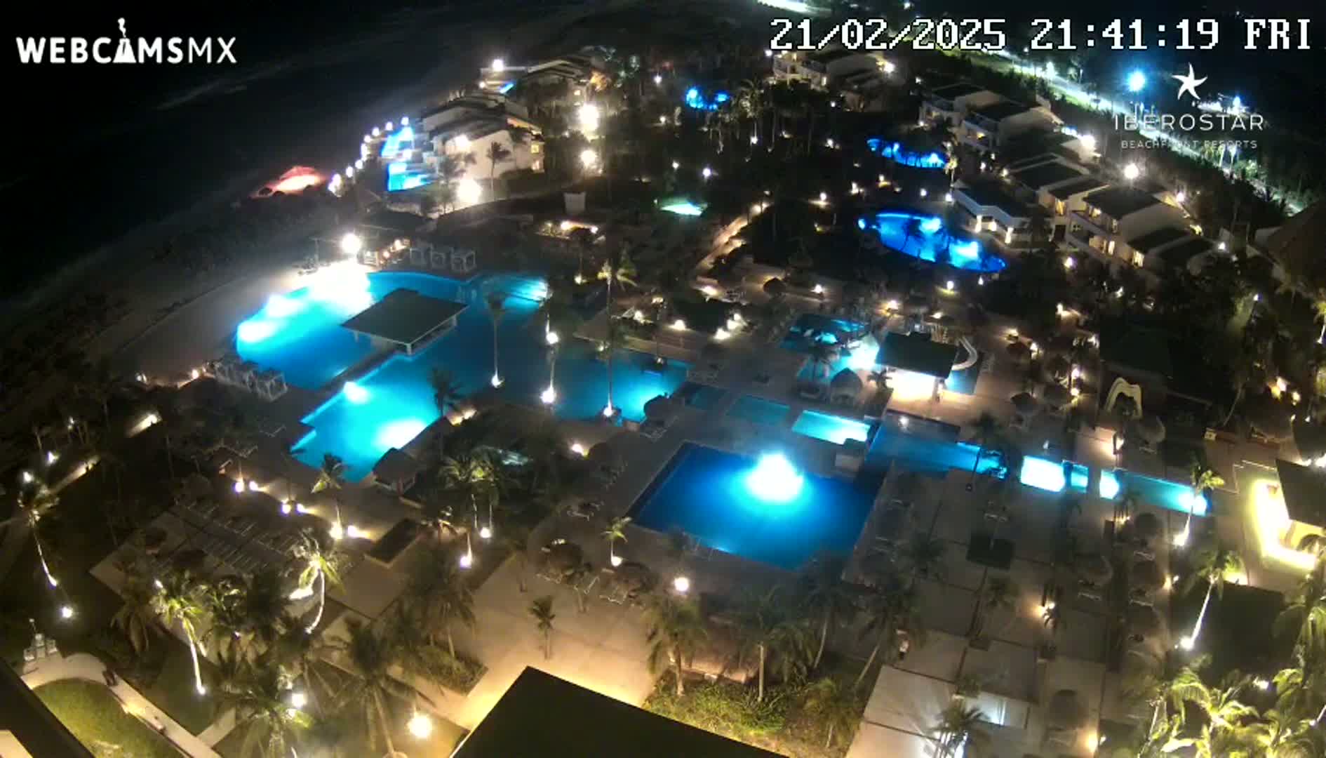 An aerial nighttime view of a resort complex with numerous illuminated pools and surrounding buildings, next to a dark ocean.