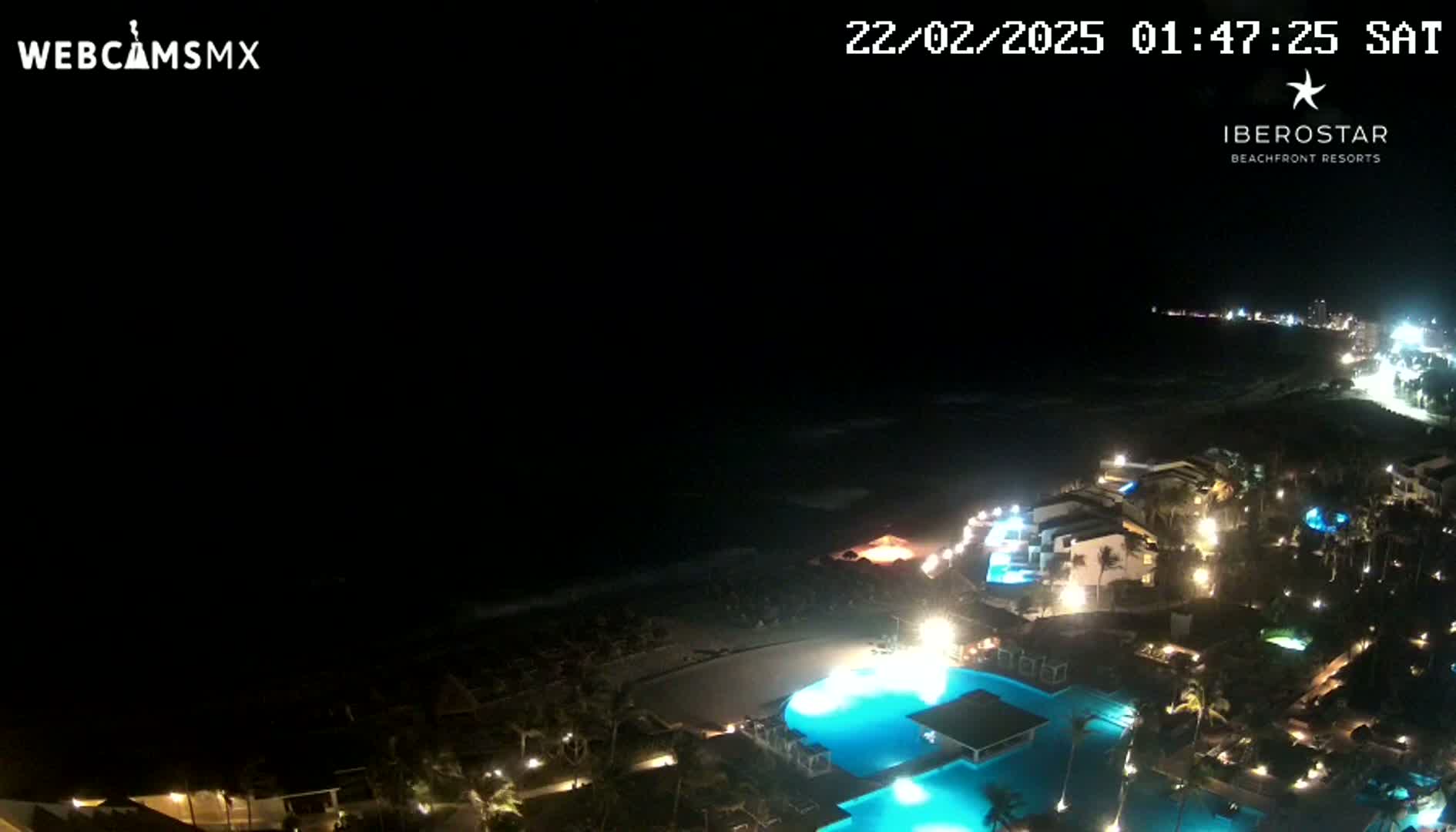 A nighttime aerial view of a resort complex illuminated along a dark ocean shore under a clear sky.