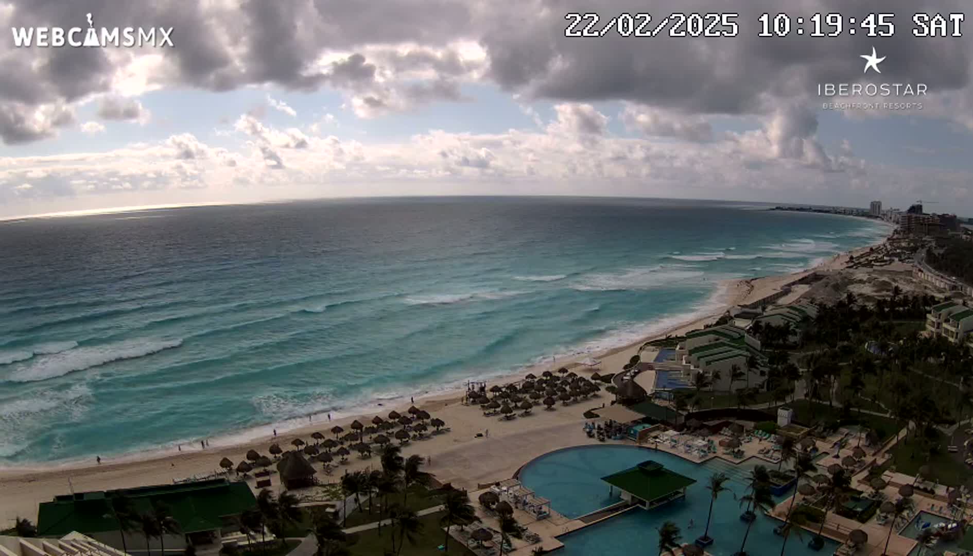 An ocean beach with turquoise water and moderate waves is partially visible under a partly cloudy sky, with a resort and swimming pools situated along the shore.