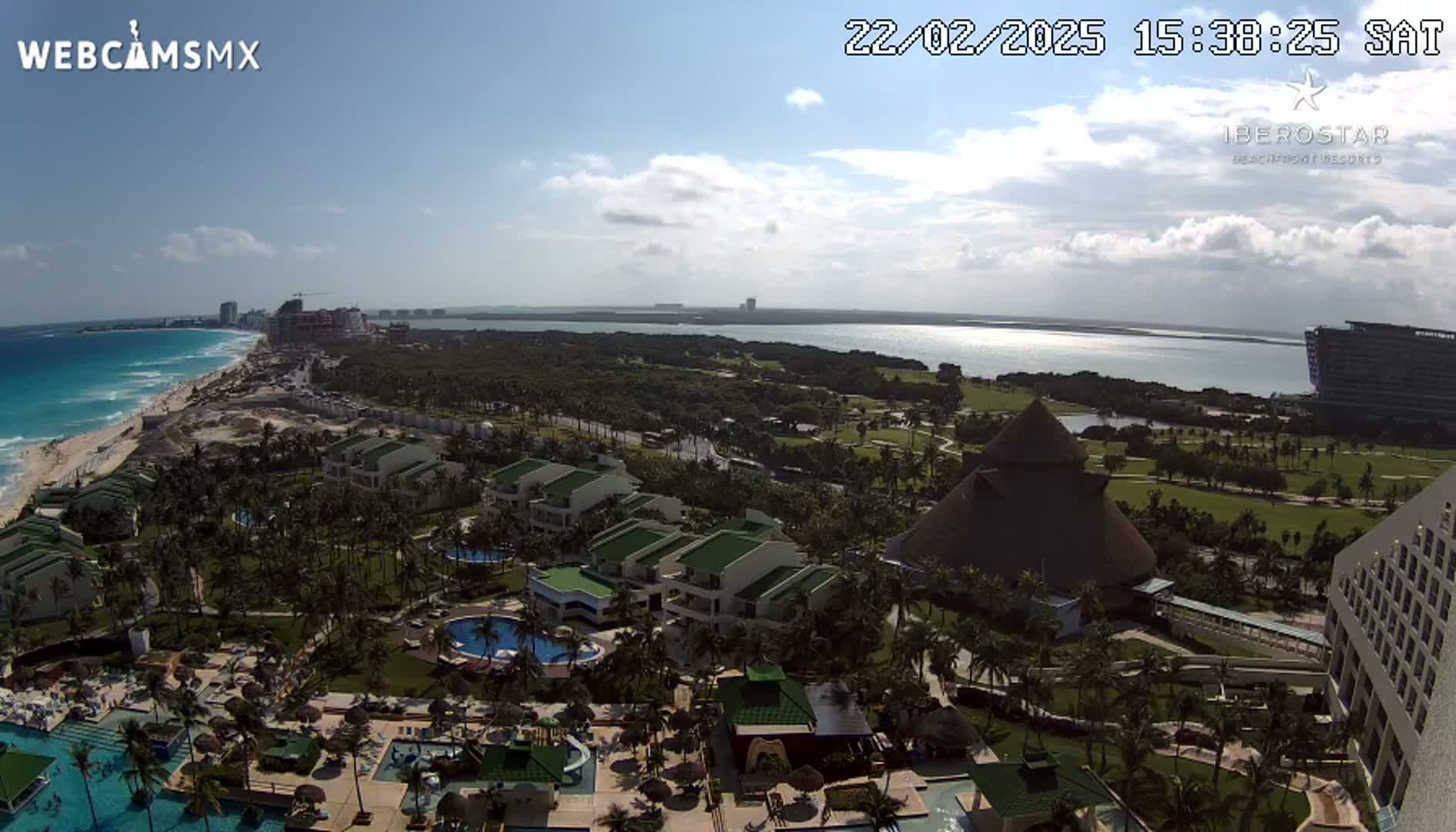An aerial view shows a resort complex with pools and buildings situated on a tropical coast next to a calm ocean under a partly cloudy sky.