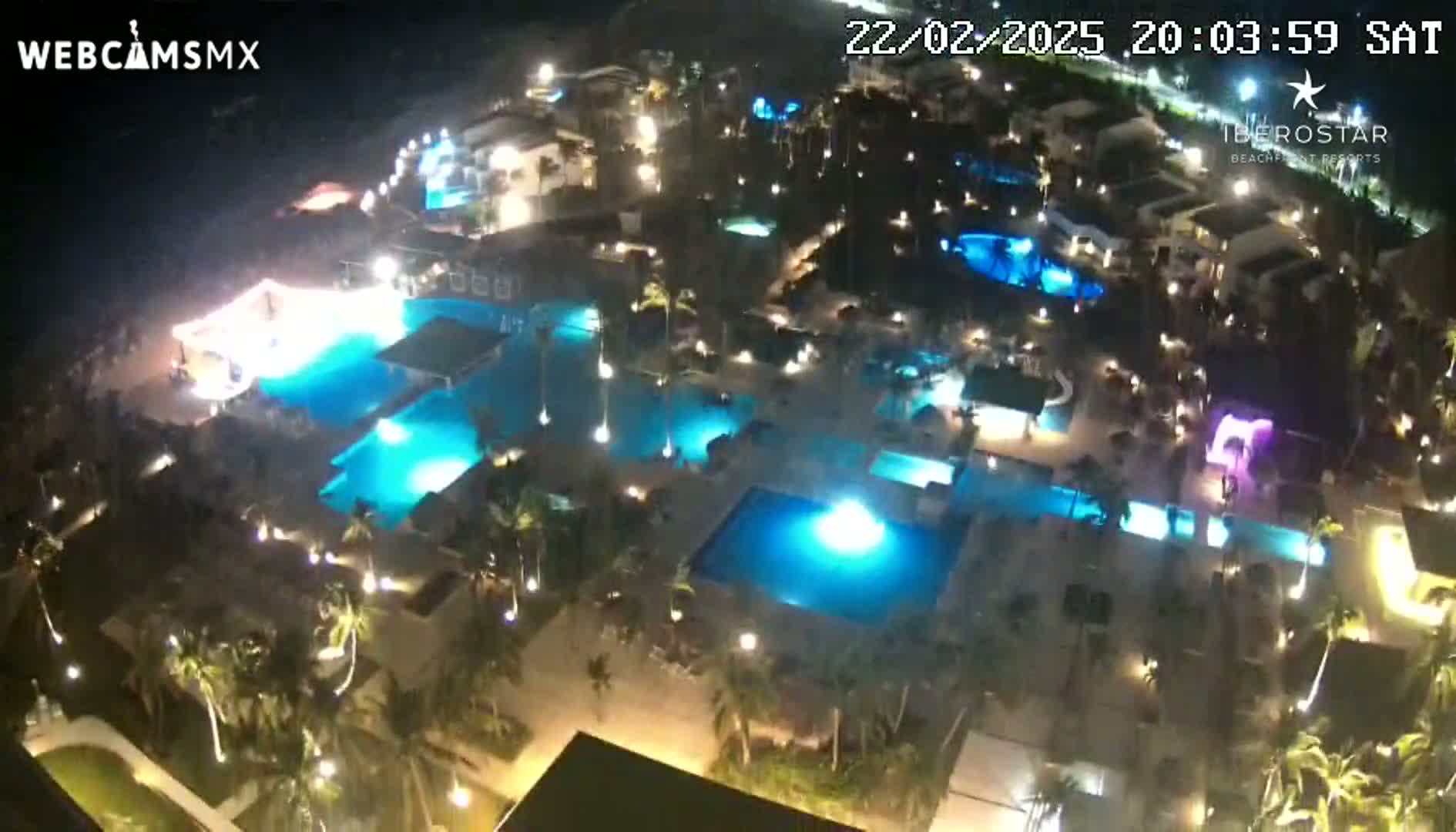 An aerial nighttime view shows a resort complex with numerous illuminated swimming pools surrounded by palm trees and buildings.