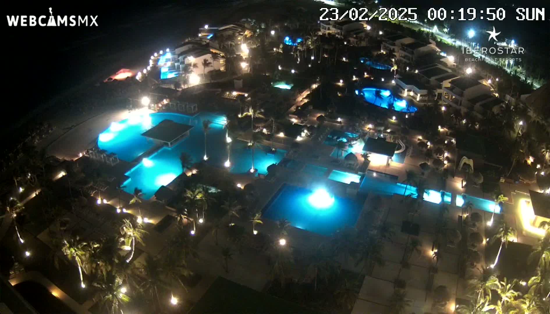 An aerial nighttime view shows a resort complex with numerous illuminated swimming pools surrounded by palm trees and buildings.