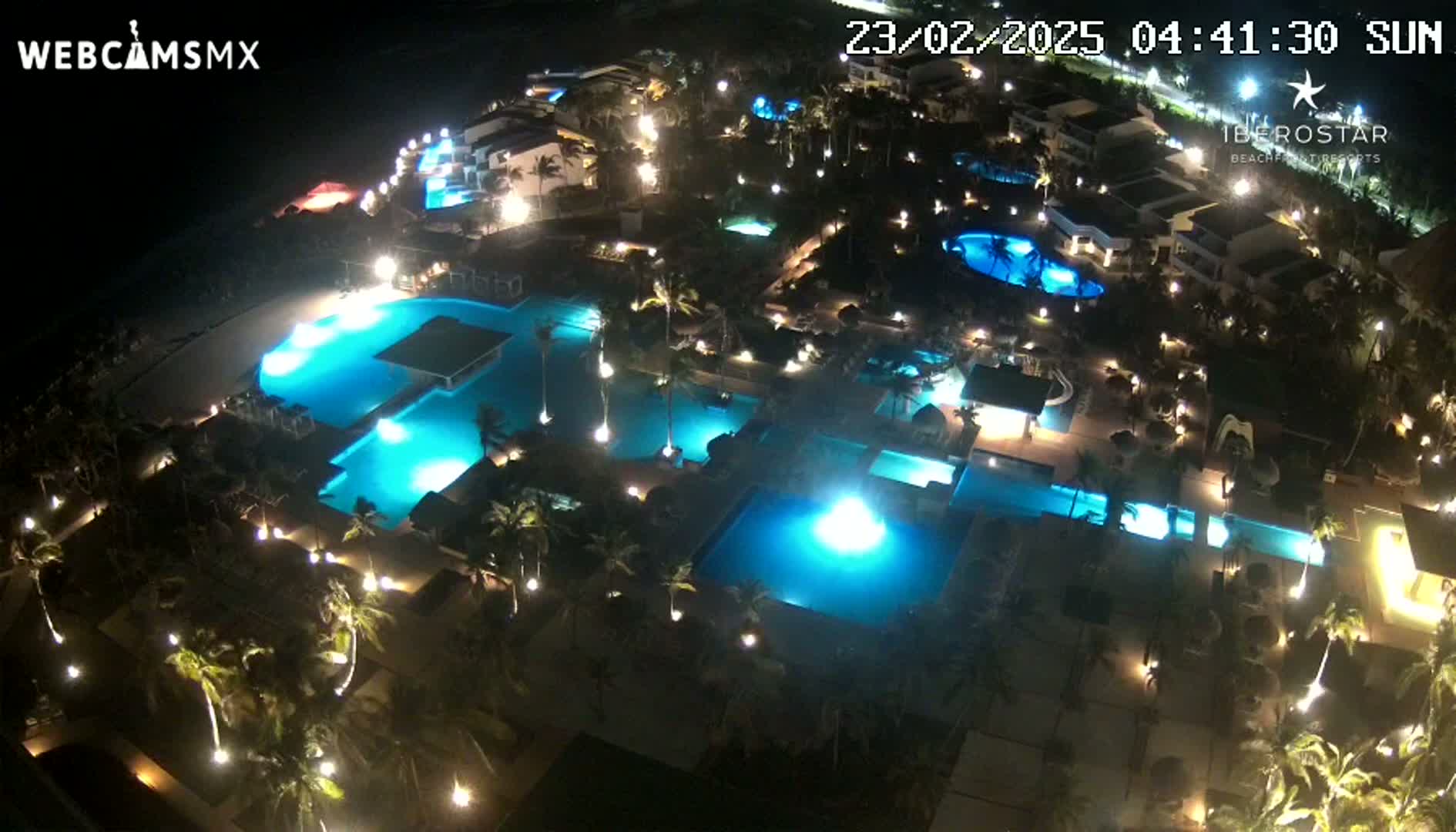An aerial nighttime view of a resort complex with numerous illuminated swimming pools surrounded by palm trees and buildings.