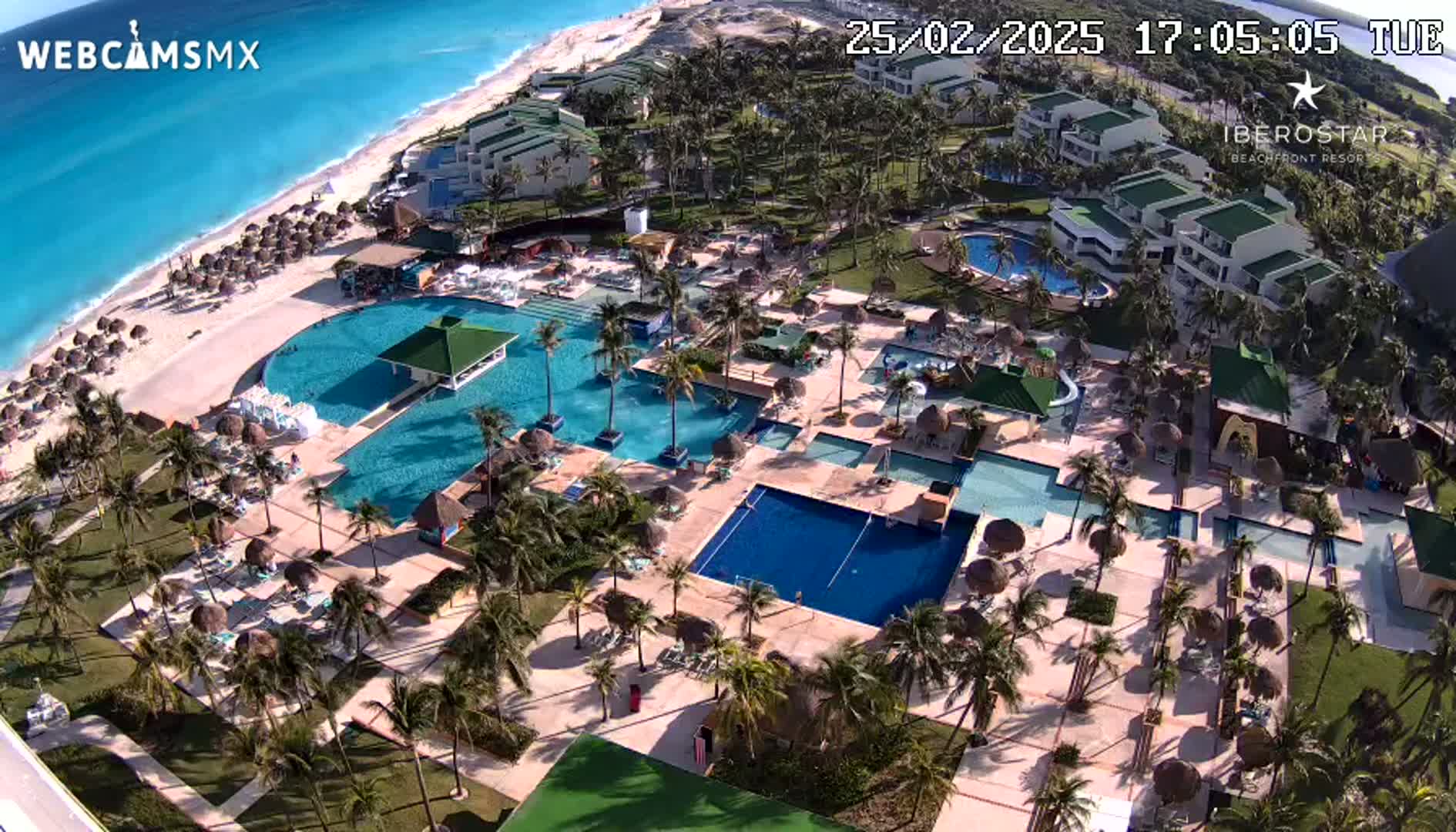 An aerial view of a resort with numerous pools, beach access, and palm trees on a sunny day.