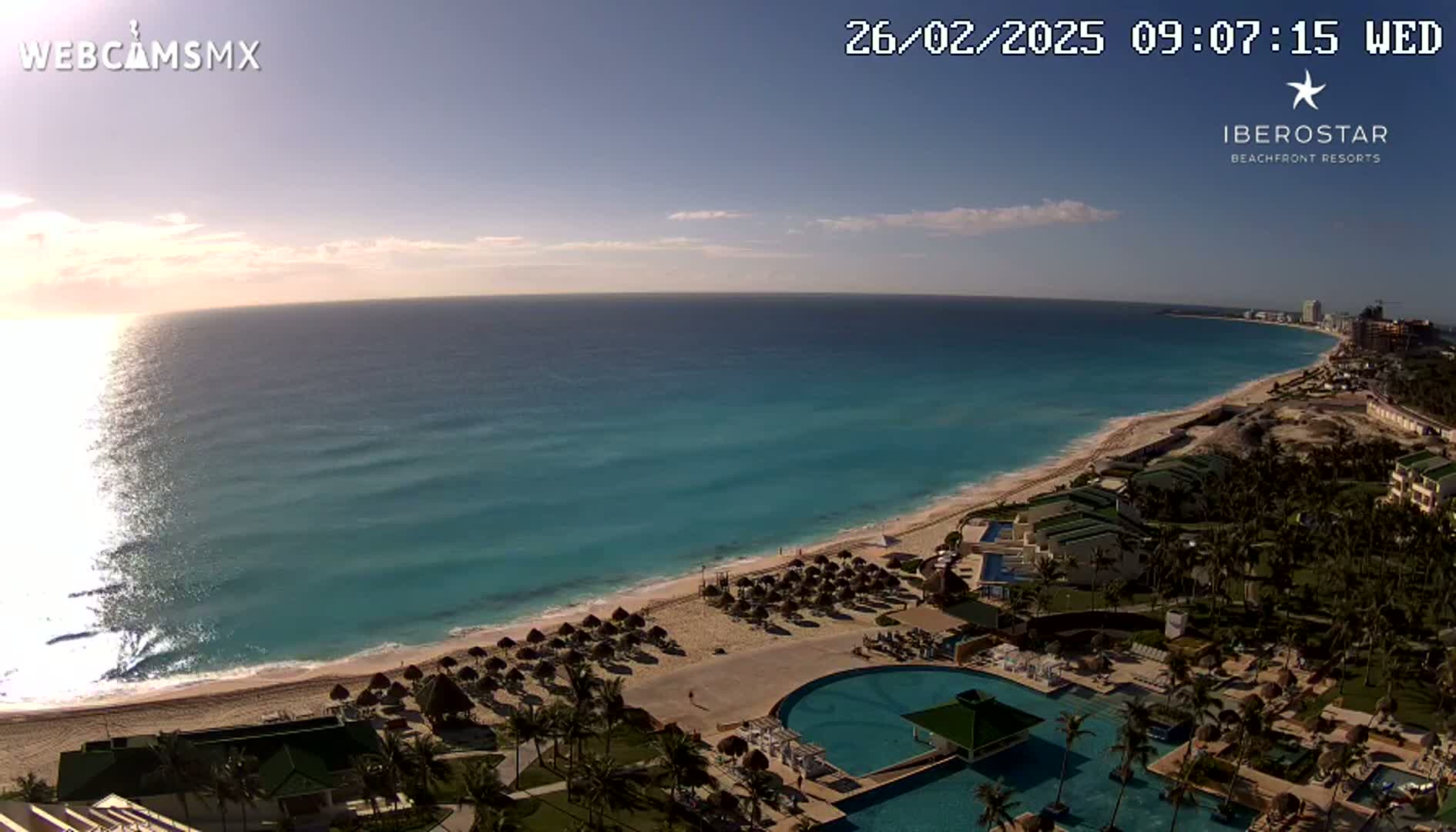 A beach resort with a large pool sits on a sandy beach next to a turquoise ocean under a mostly clear morning sky.