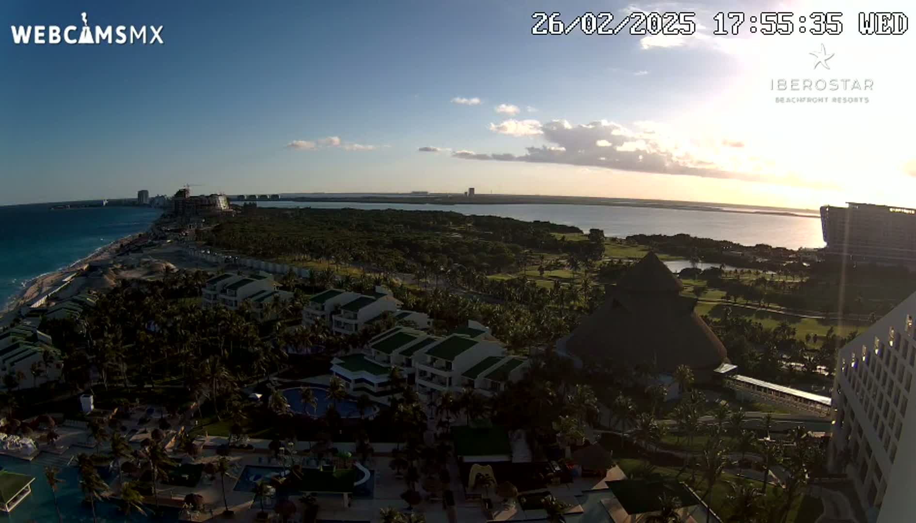 An aerial view of a tropical resort at sunset, showing buildings, pools, lush vegetation, and a body of water beyond.
