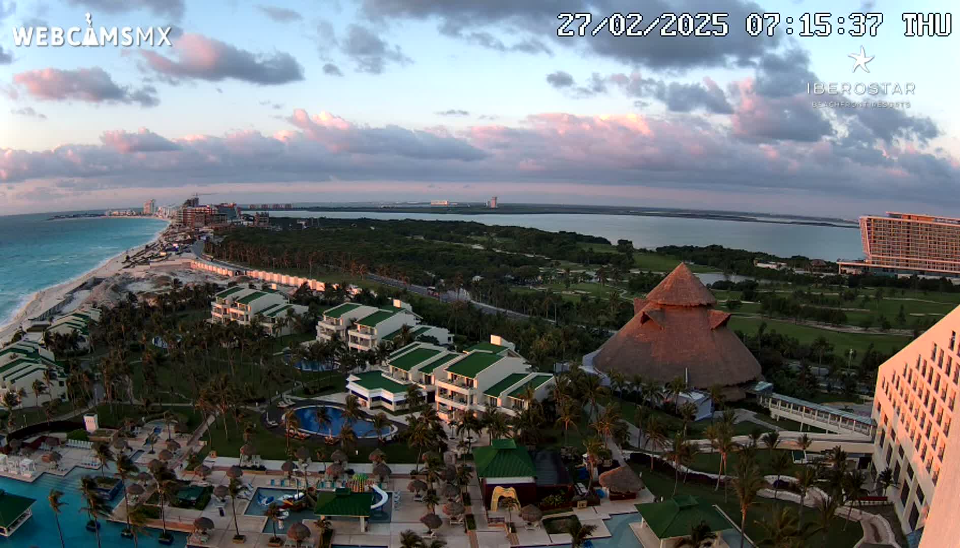 An aerial view of a resort at sunrise, showing low-lying clouds, a beach, ocean, and various buildings with pools and palm trees.