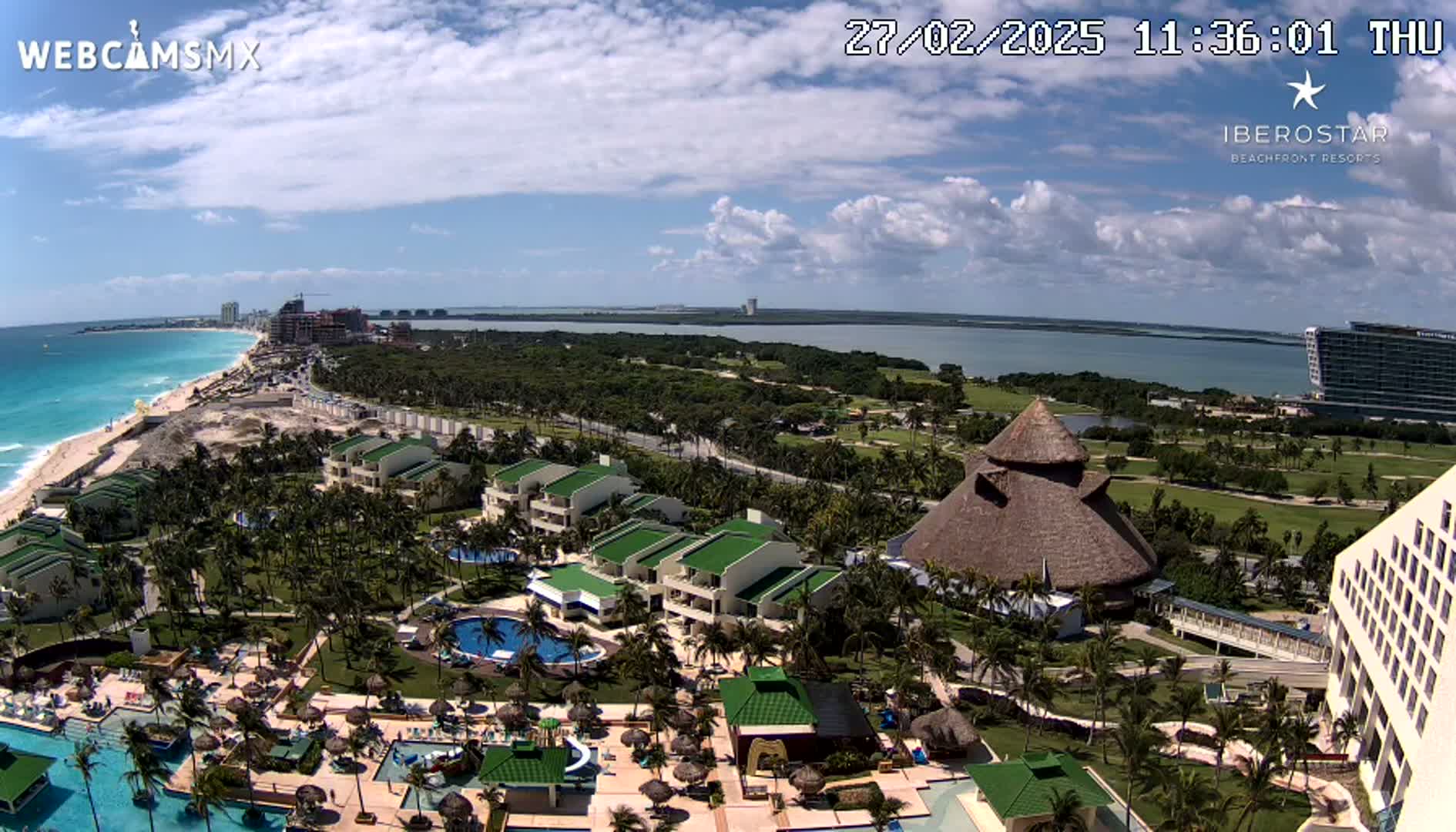 An aerial view of a resort complex on a sunny day, featuring turquoise ocean water, palm trees, pools, and buildings, with a calm body of water visible in the distance.