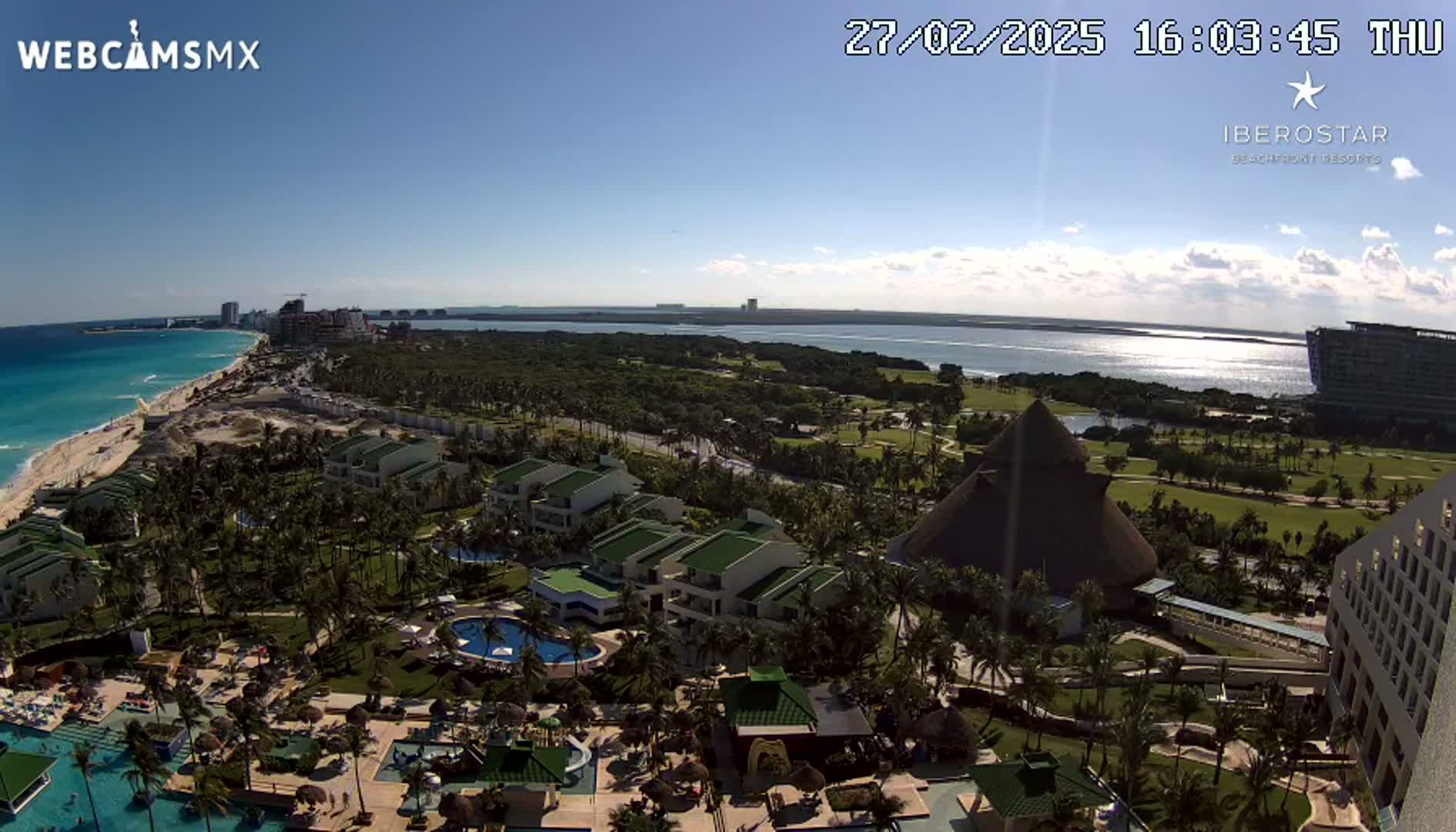 A sunny, aerial view of a resort town situated on a coastline with a turquoise ocean, featuring lush greenery, buildings, pools, and a large thatched structure.