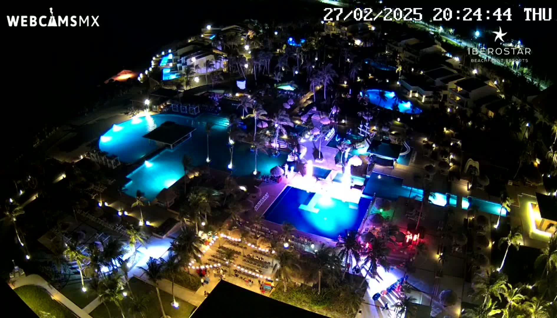 An aerial nighttime view of a resort with brightly lit pools and palm trees, under a clear sky.