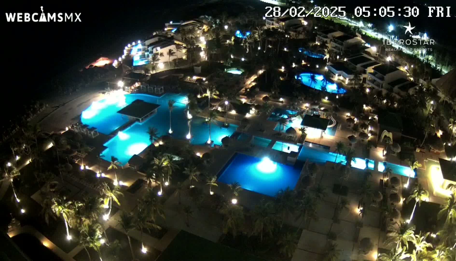 An illuminated resort at night, featuring numerous swimming pools surrounded by palm trees and buildings.