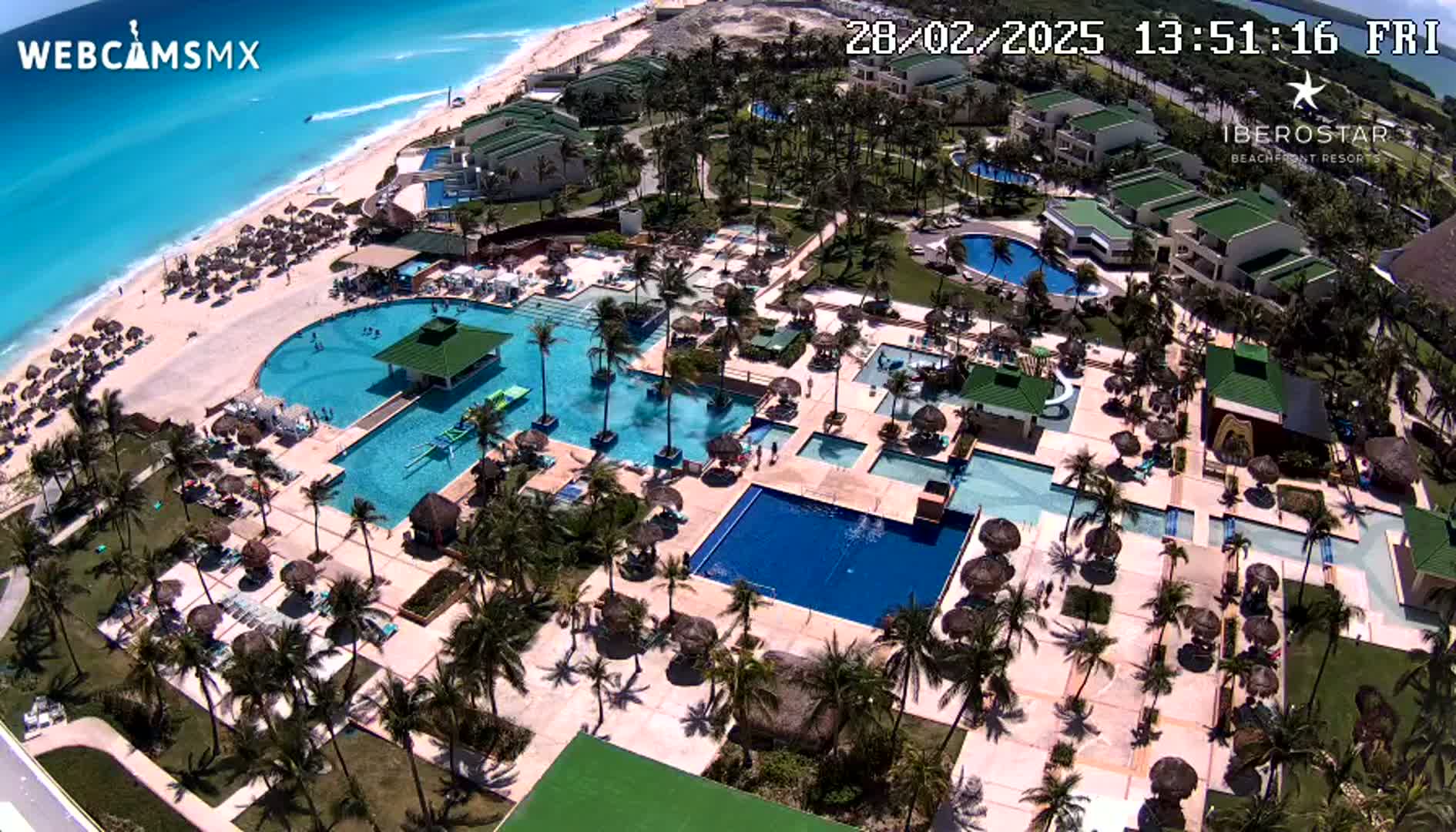 An aerial view of a resort on a sunny day shows a large complex of pools, a sandy beach, and turquoise ocean.