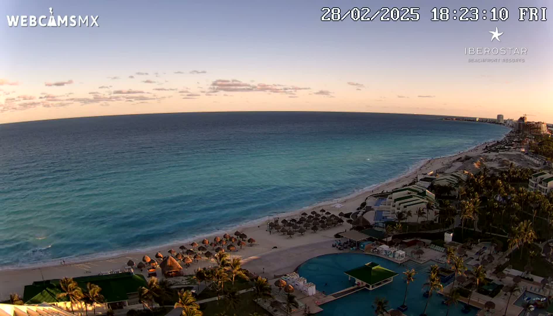 A beach resort at sunset overlooks a calm, turquoise ocean under a mostly clear sky.