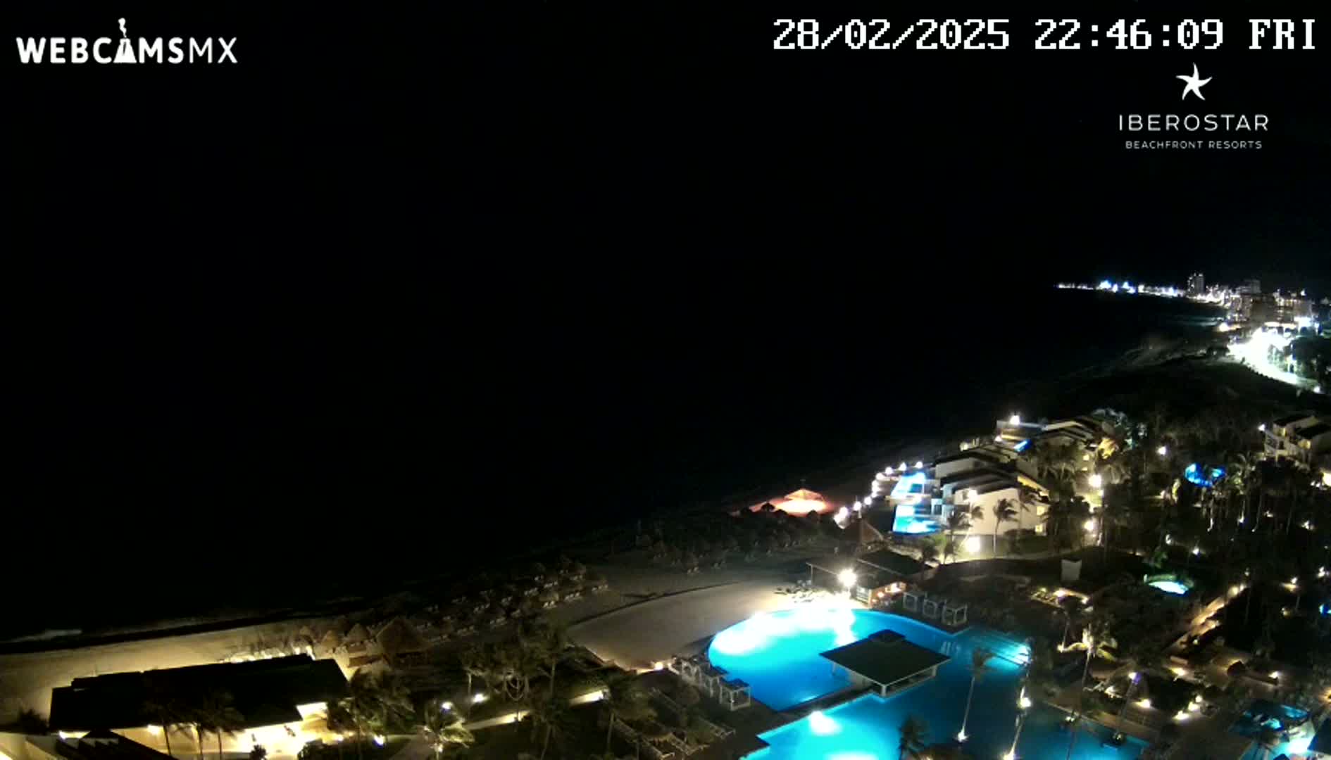 A nighttime, aerial view of a coastal resort complex with illuminated pools and buildings next to a dark ocean under a clear, dark sky.