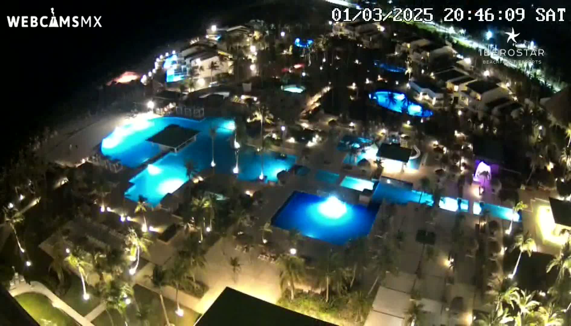 A nighttime aerial view shows a resort complex with numerous illuminated swimming pools surrounded by palm trees and buildings.