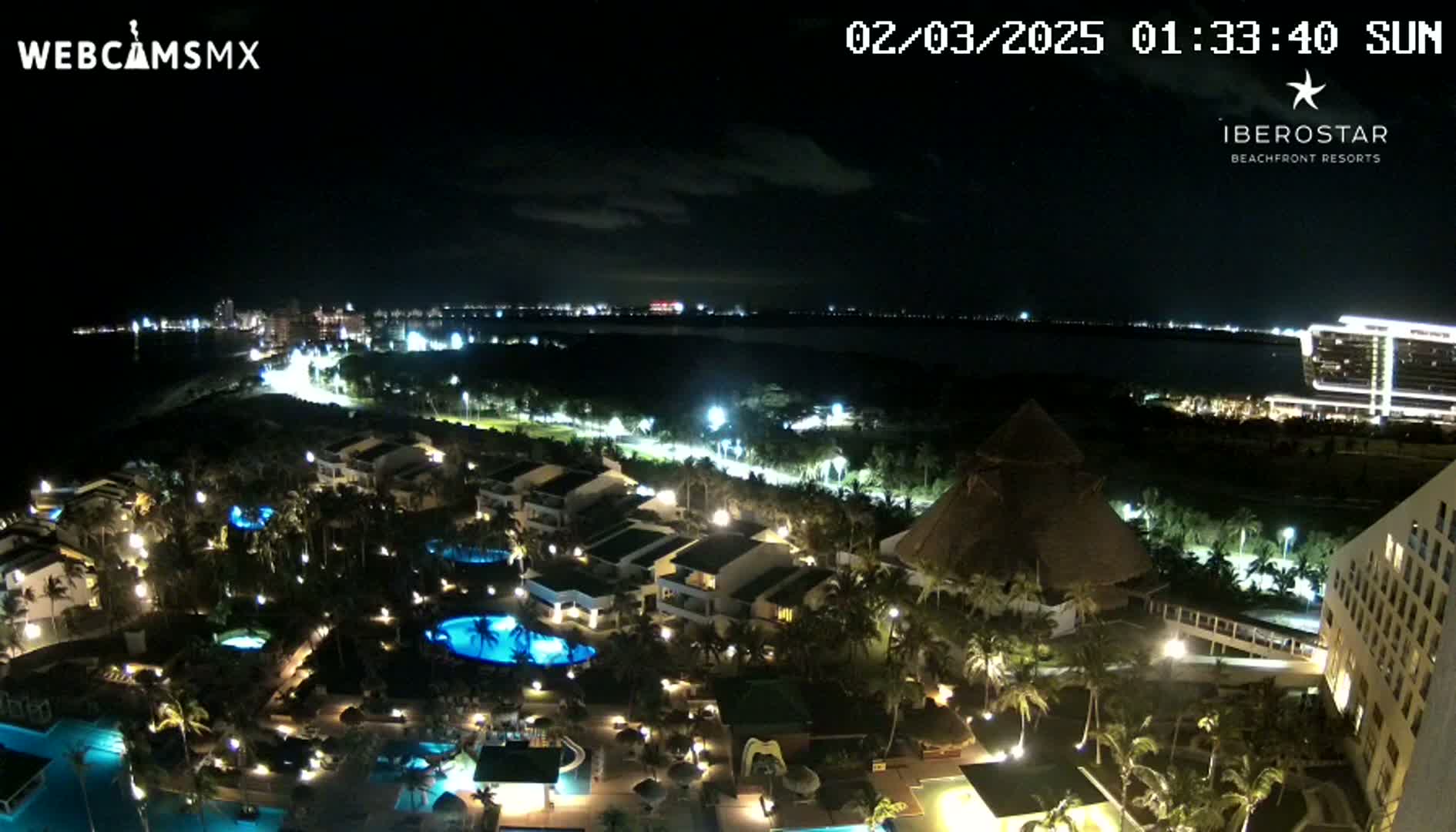 A nighttime aerial view of a resort complex with pools and palm trees, situated near a body of water with a city skyline visible in the distance under a clear sky.