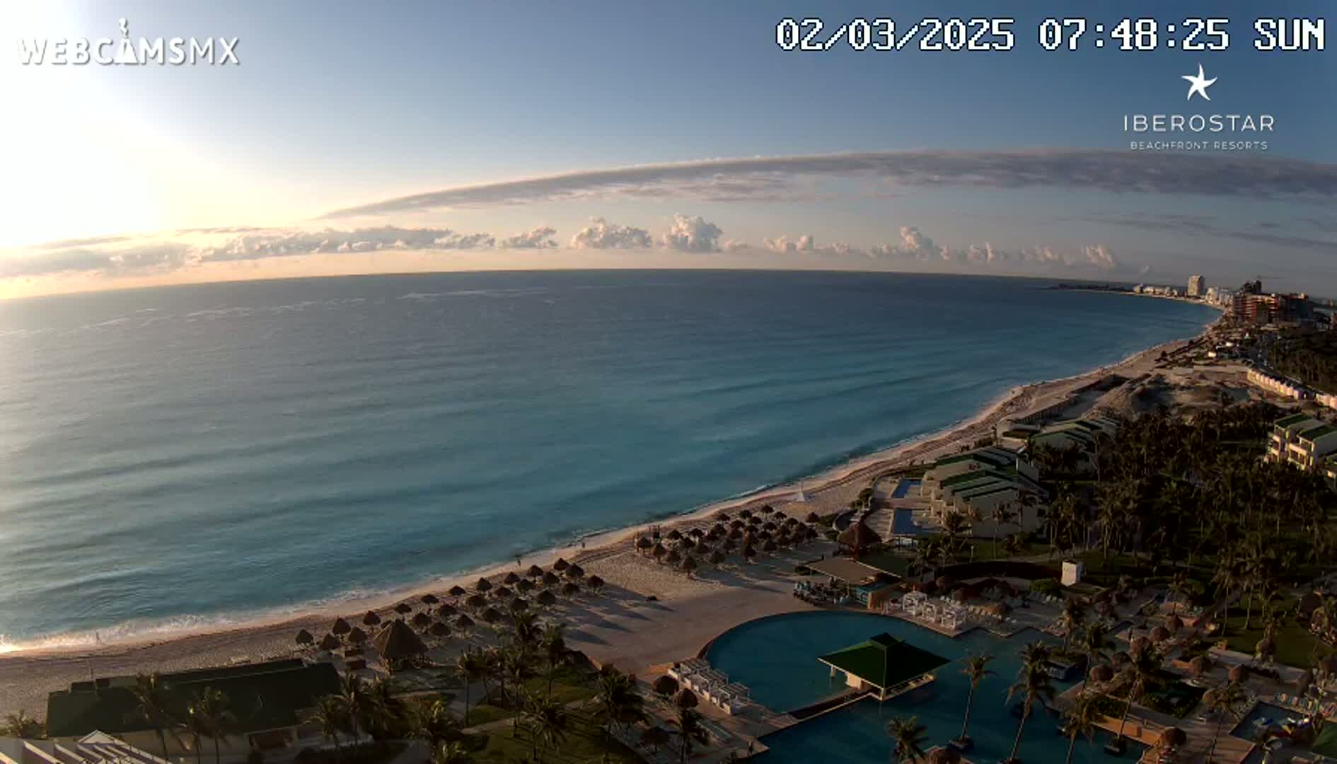 A sunrise view shows a long sandy beach with turquoise water, numerous palm trees, and a resort with pools under a mostly clear sky.